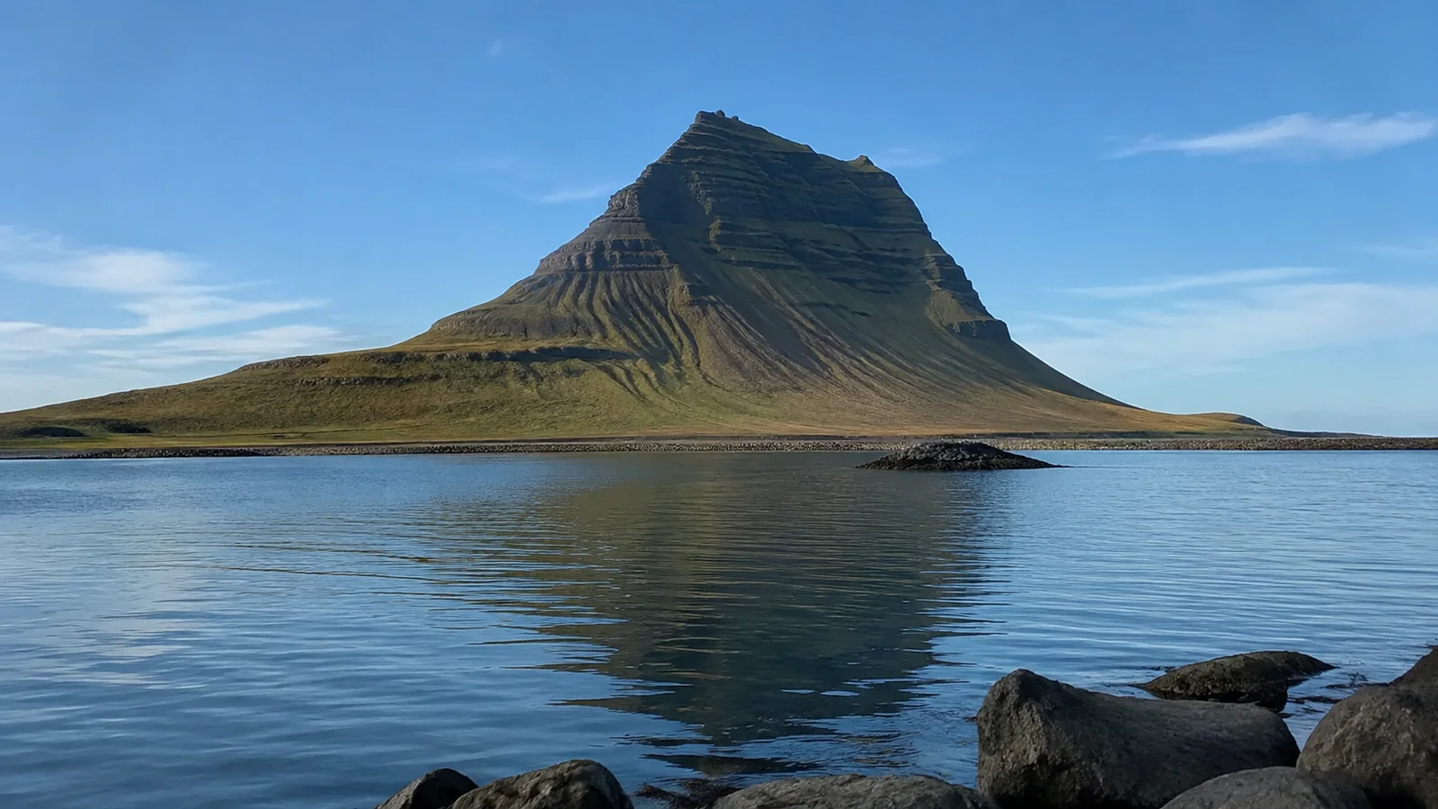 Steep isolated mountain reflected in calm coastal water under blue skies.
