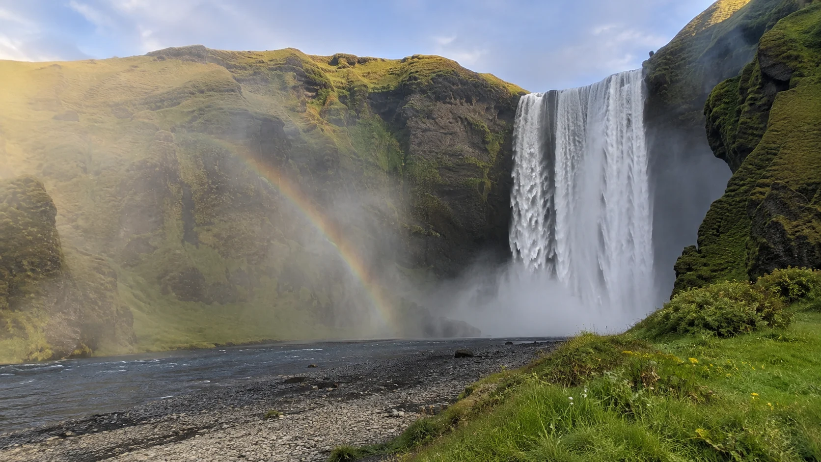 Skógafoss cascading from a green cliff with a rainbow in its mist.