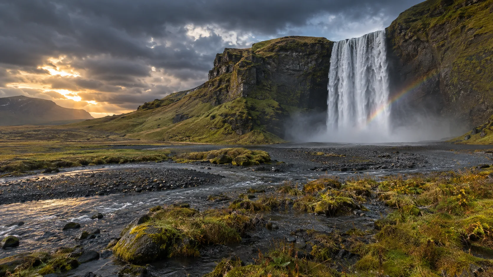 Skógafoss waterfall, black volcanic gravel and rainbow spray beneath stormy Icelandic skies