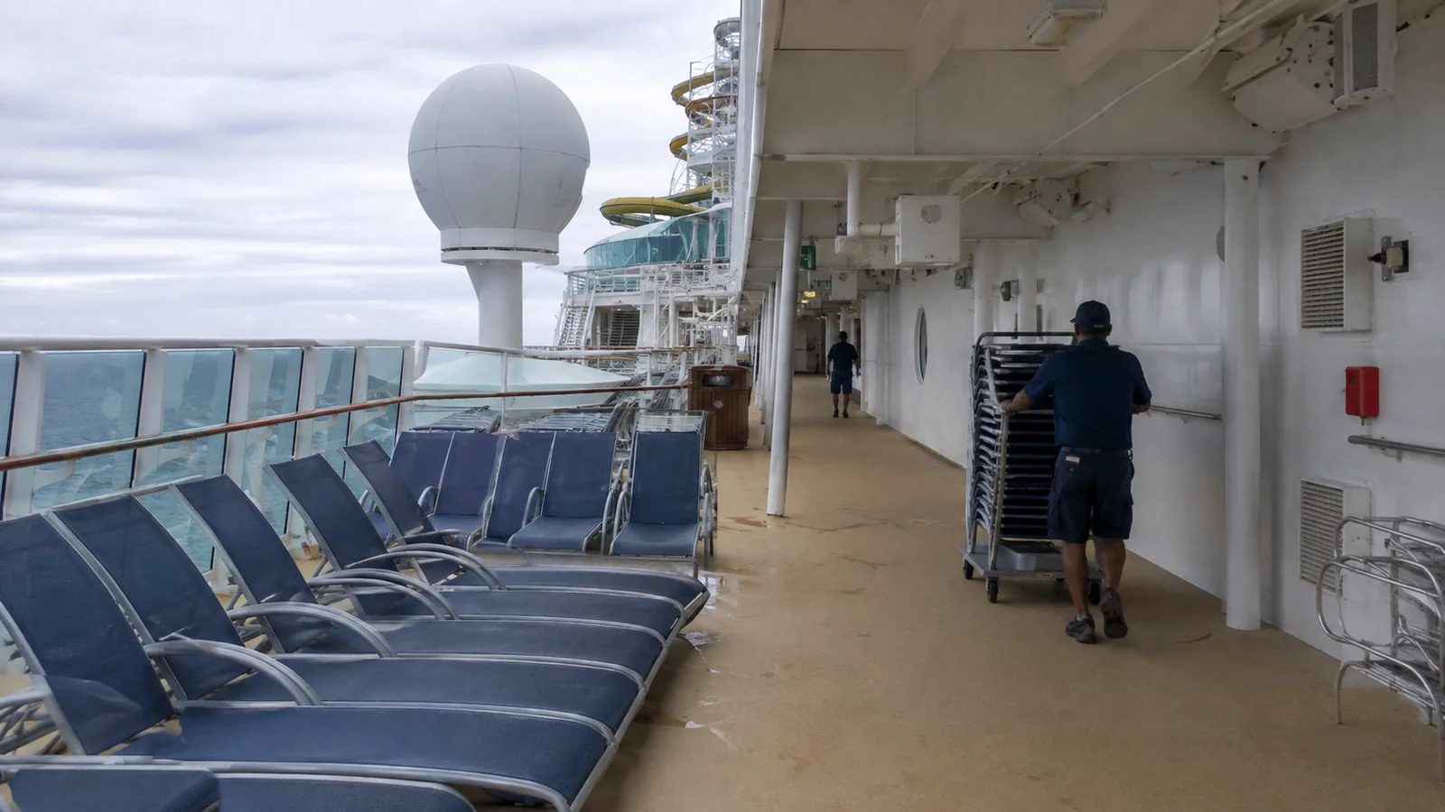 Blue lounge chairs along a cruise ship promenade with a worker moving a cart