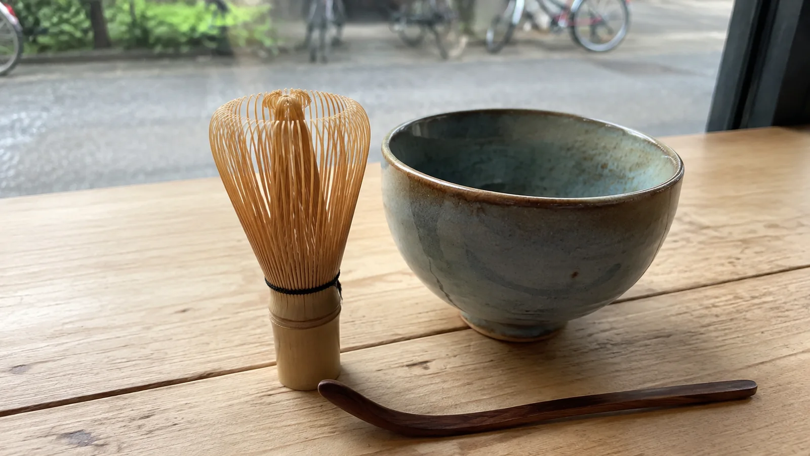 Bamboo whisk and ceramic bowl on a wooden surface beside a window.