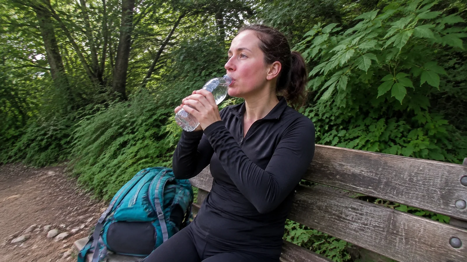 Woman drinking water on a forest bench beside her backpack.