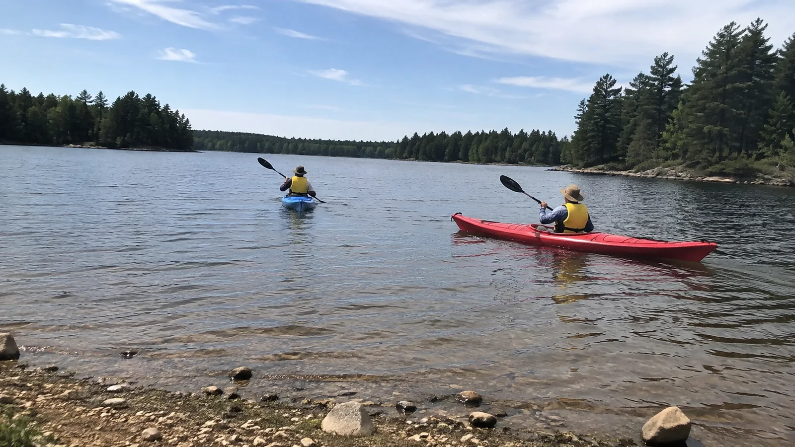 Two kayakers paddle across a lake bordered by trees and rocks.