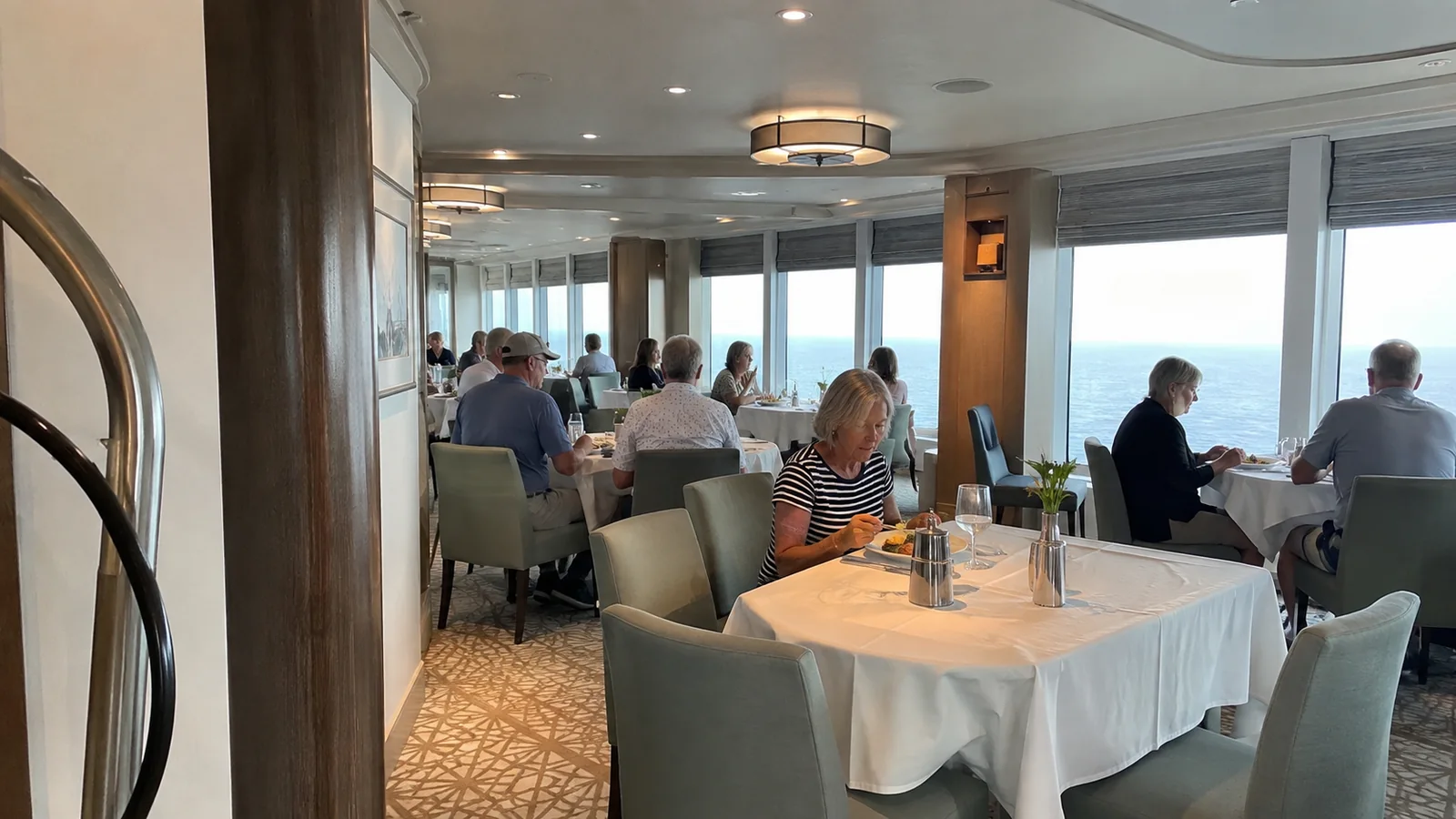 Passengers dining at white-clothed tables beside ocean-view windows.