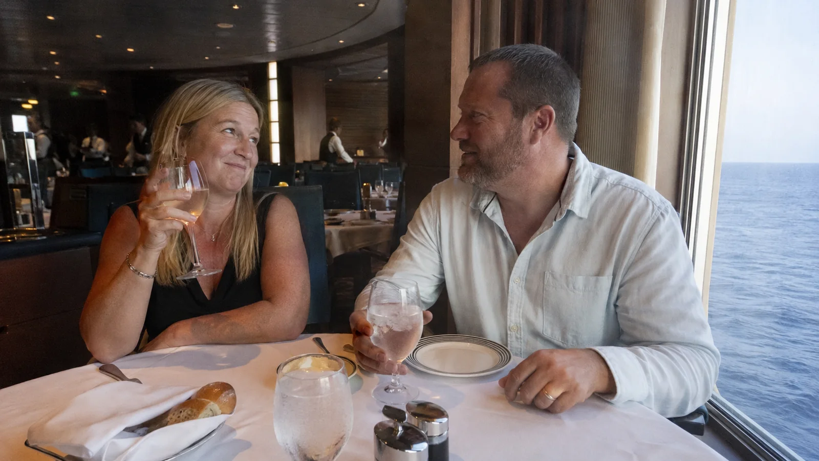 Couple holds glasses while dining beside an ocean window.