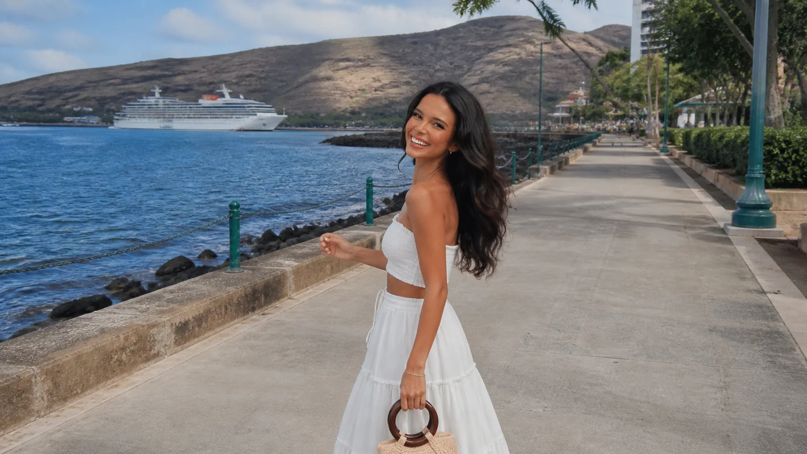 Woman in a white dress walking along a waterfront promenade near a cruise ship.