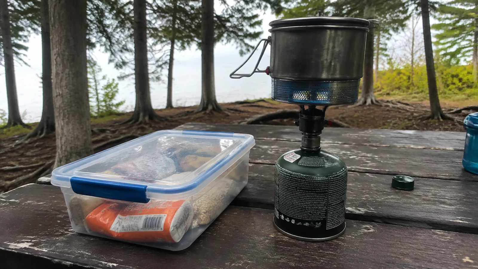 Camping stove, cooking pot, and food container on a lakeside picnic table