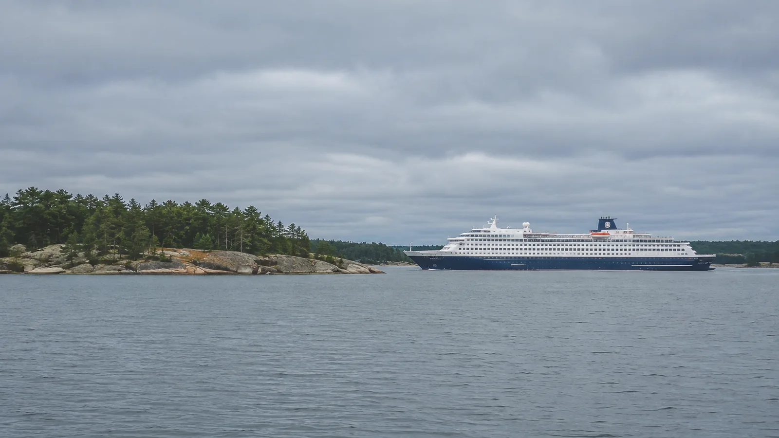 Cruise ship passing a low wooded island beneath a cloudy sky.