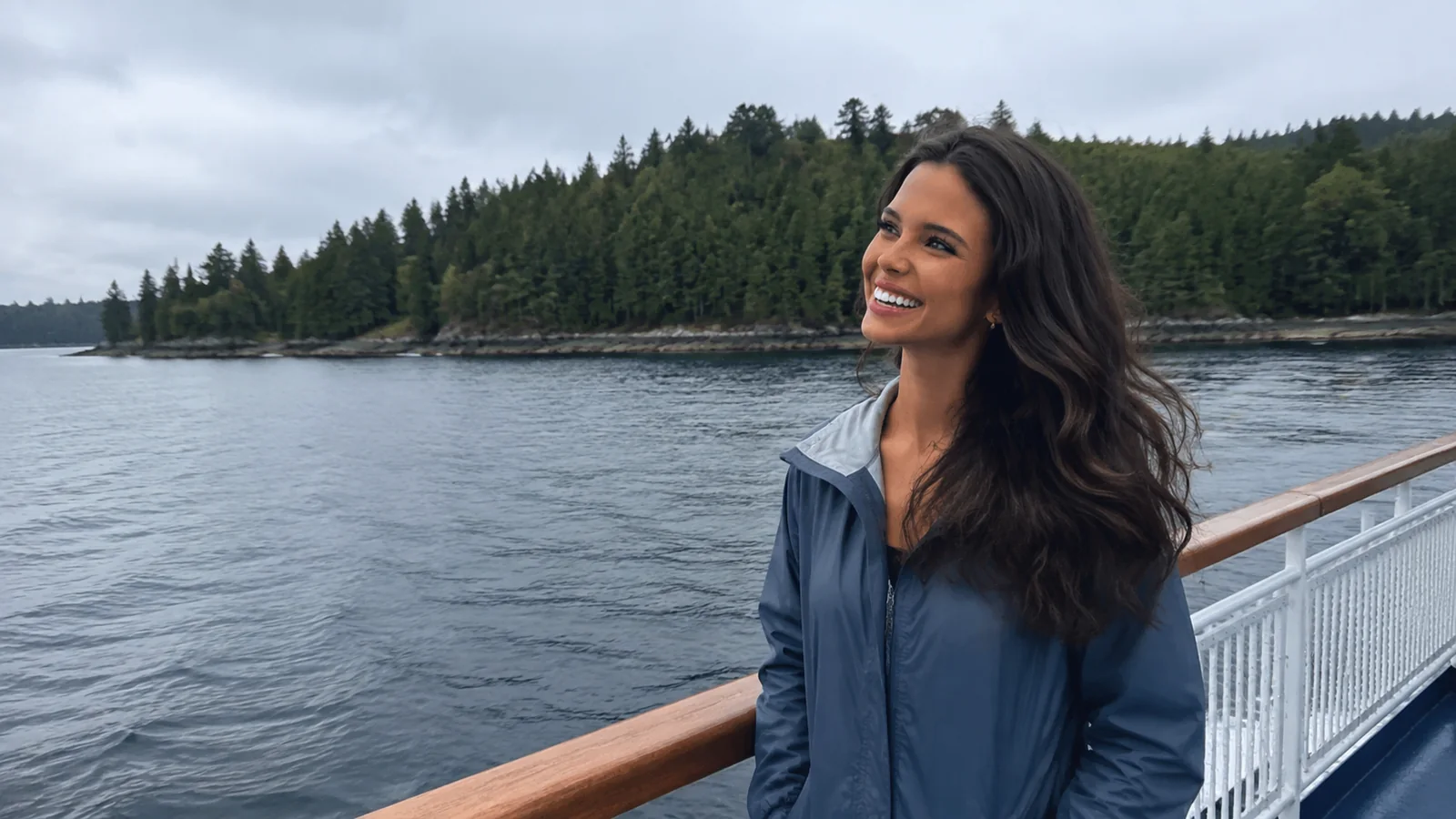 Woman in a blue jacket standing at a ship railing near a forested shoreline.