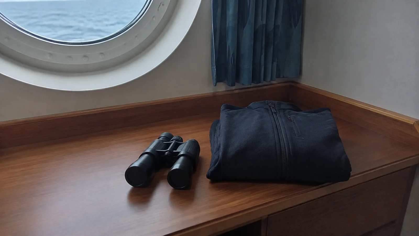 Binoculars and folded dark clothing on a wooden desk beneath a round window.
