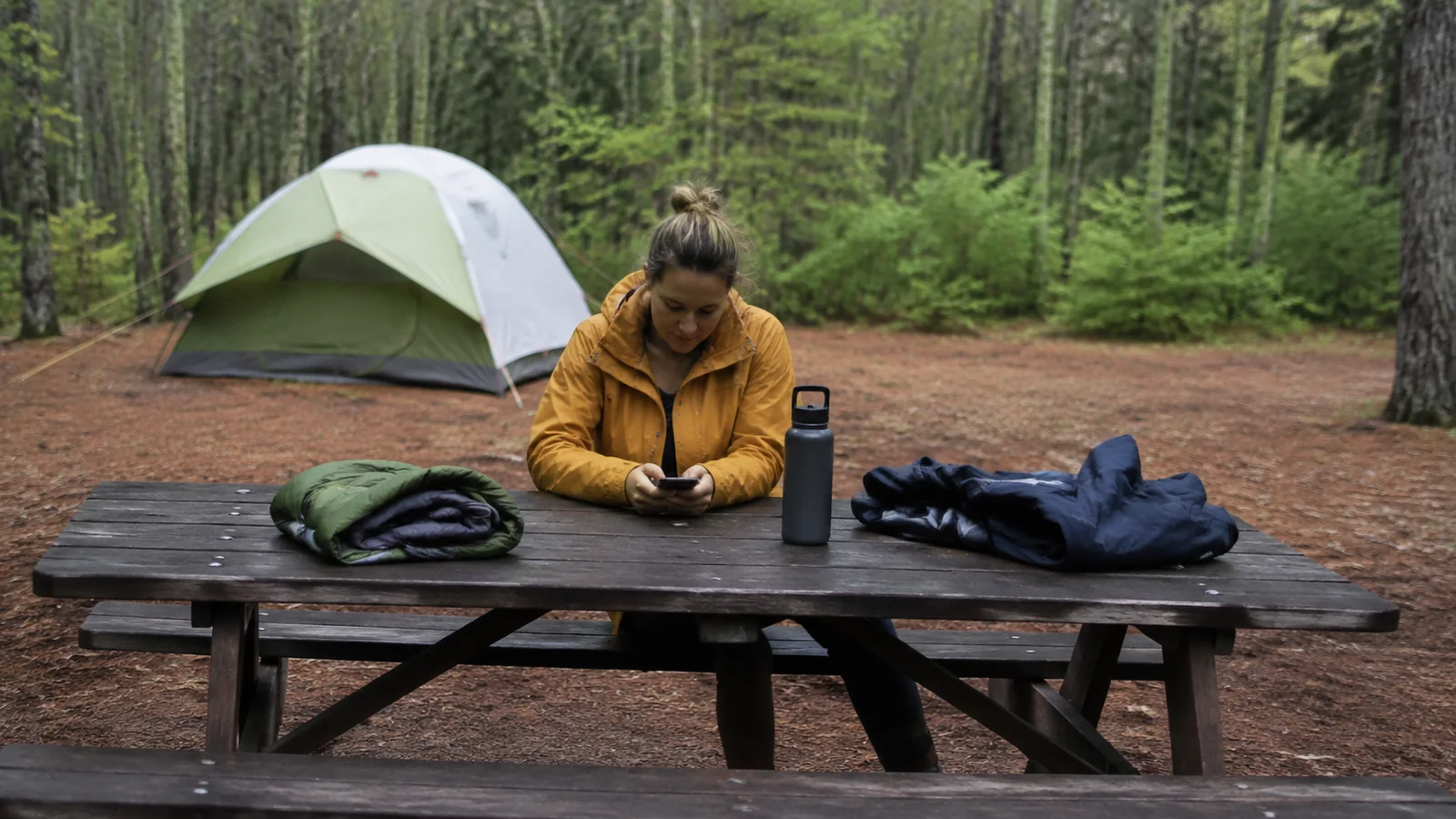 Camper seated at a picnic table with outdoor gear and a tent behind.