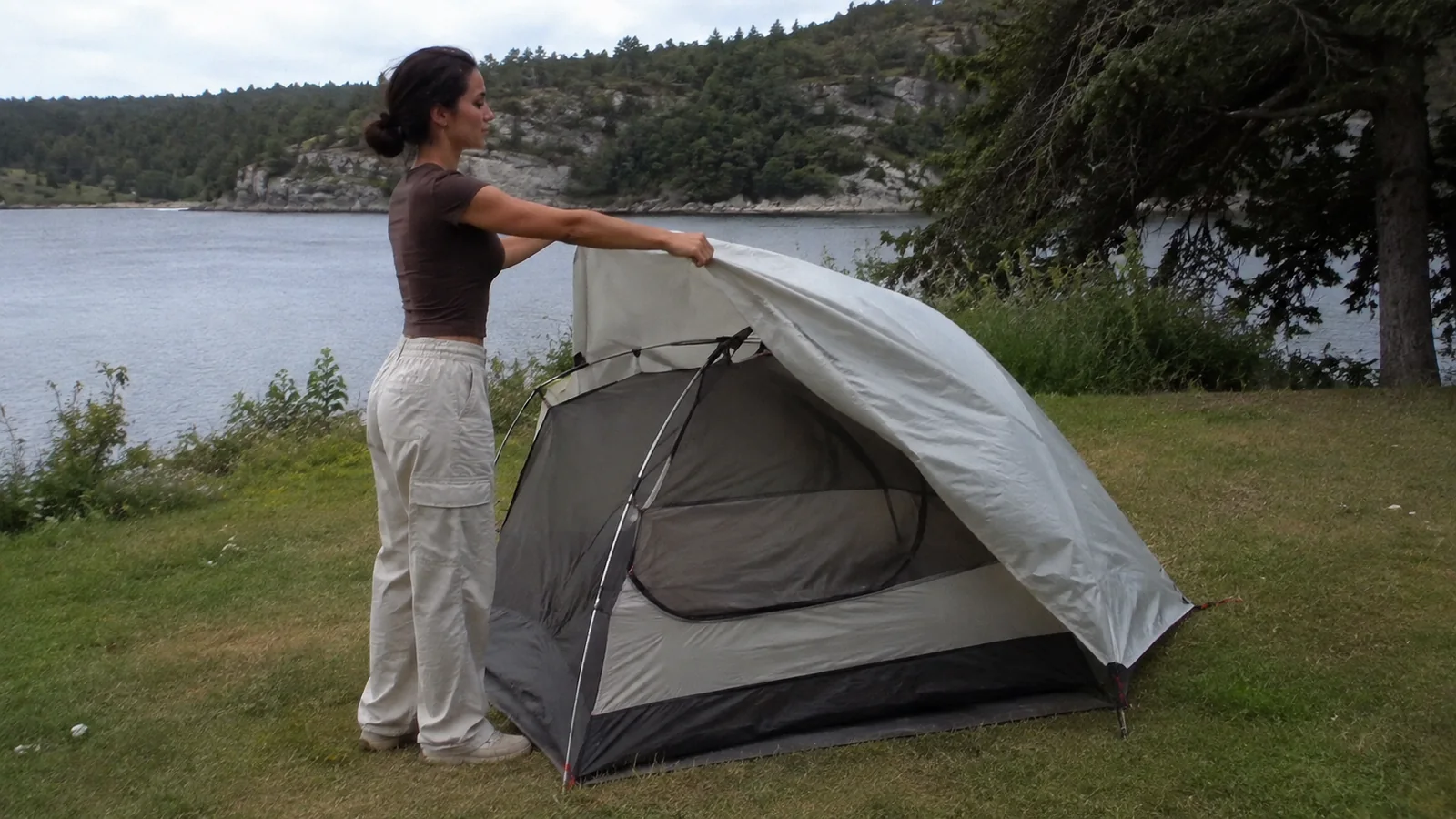 Woman adjusting a tent rainfly at a grassy lakeside campsite.