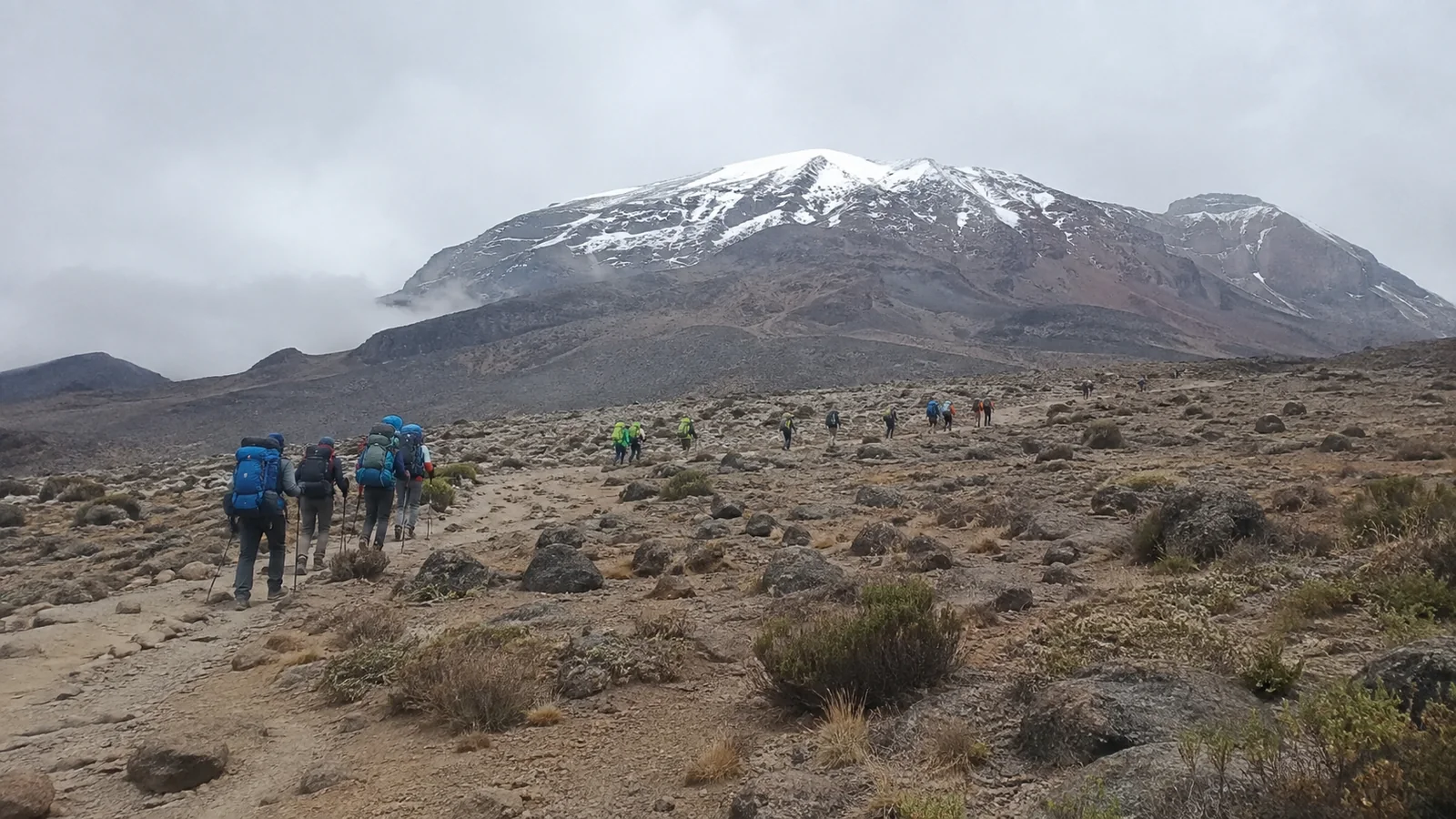 Hikers crossing rocky high-altitude terrain toward a snow-capped mountain