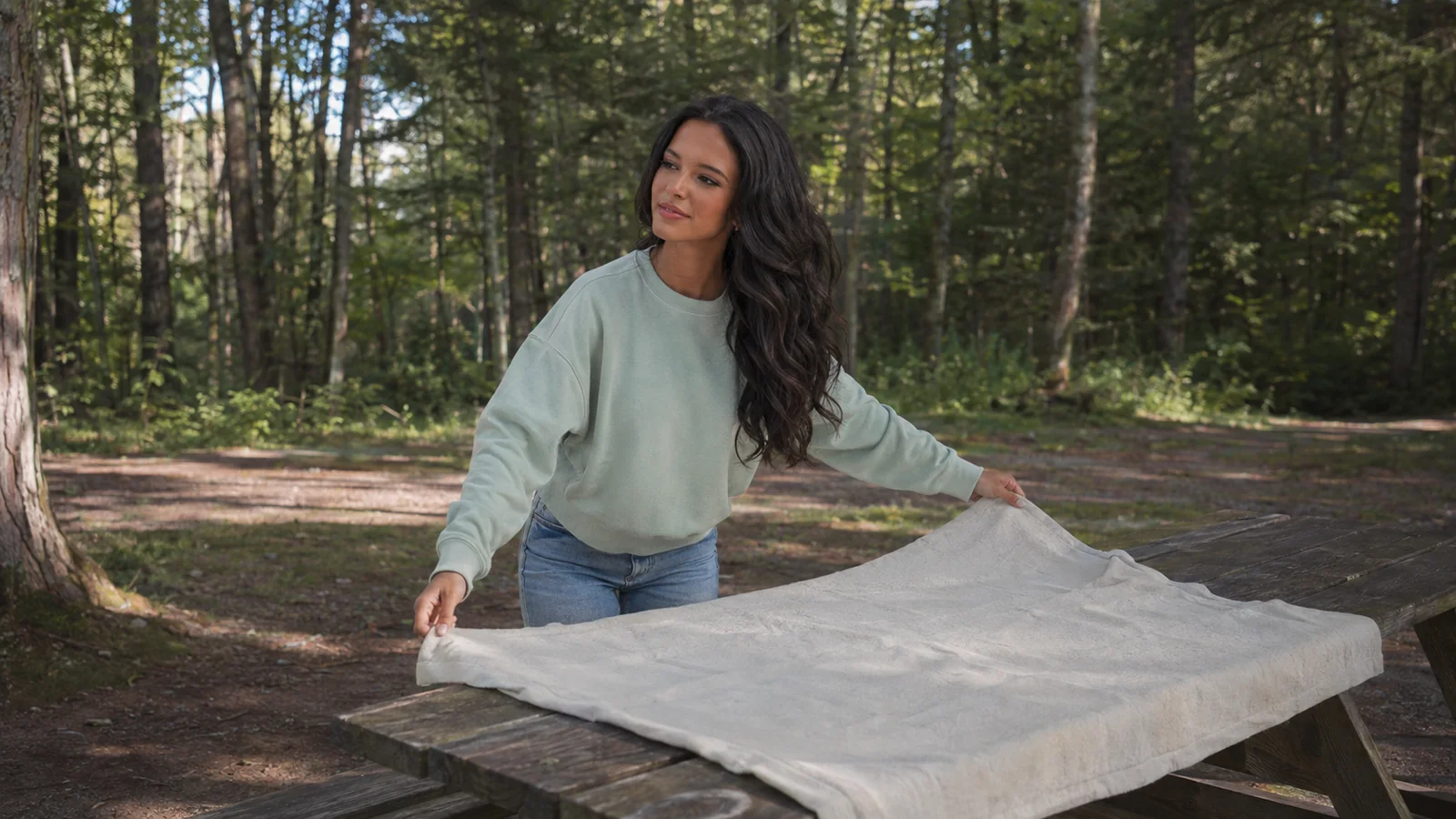 Woman spreading a cloth over a picnic table in the woods.