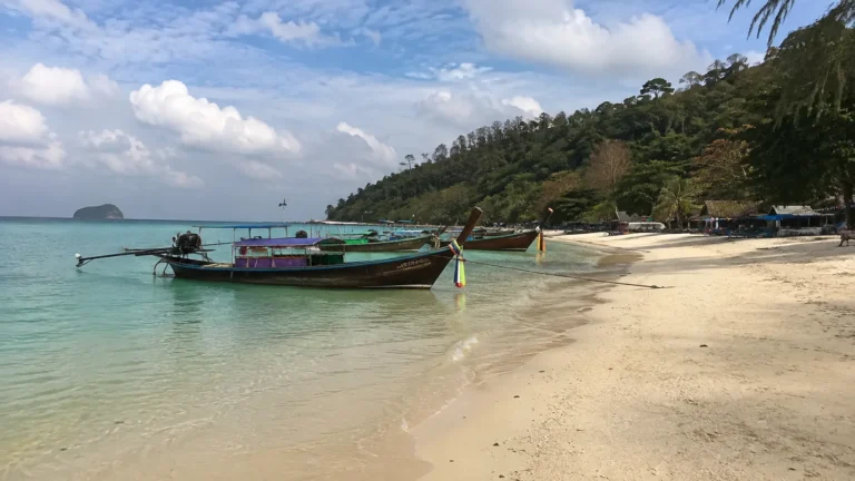Long-tail boats moored in shallow water beside a sandy tropical beach.