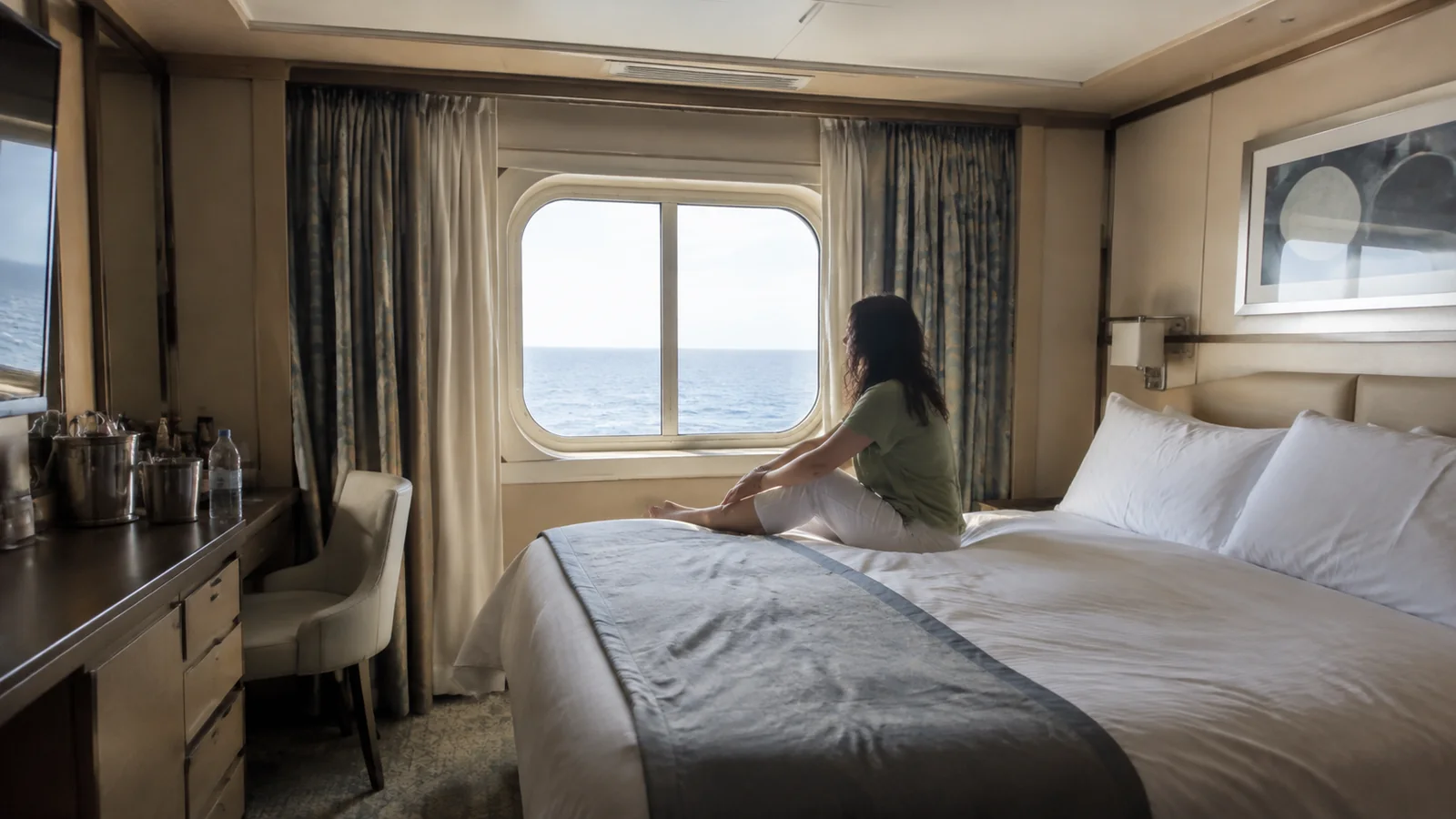 Woman sits on a stateroom bed facing an ocean window.