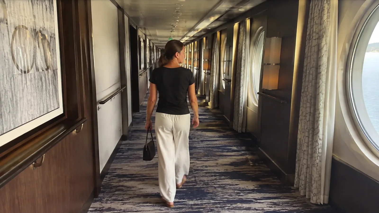 Woman walks along a carpeted ship corridor carrying a bag.