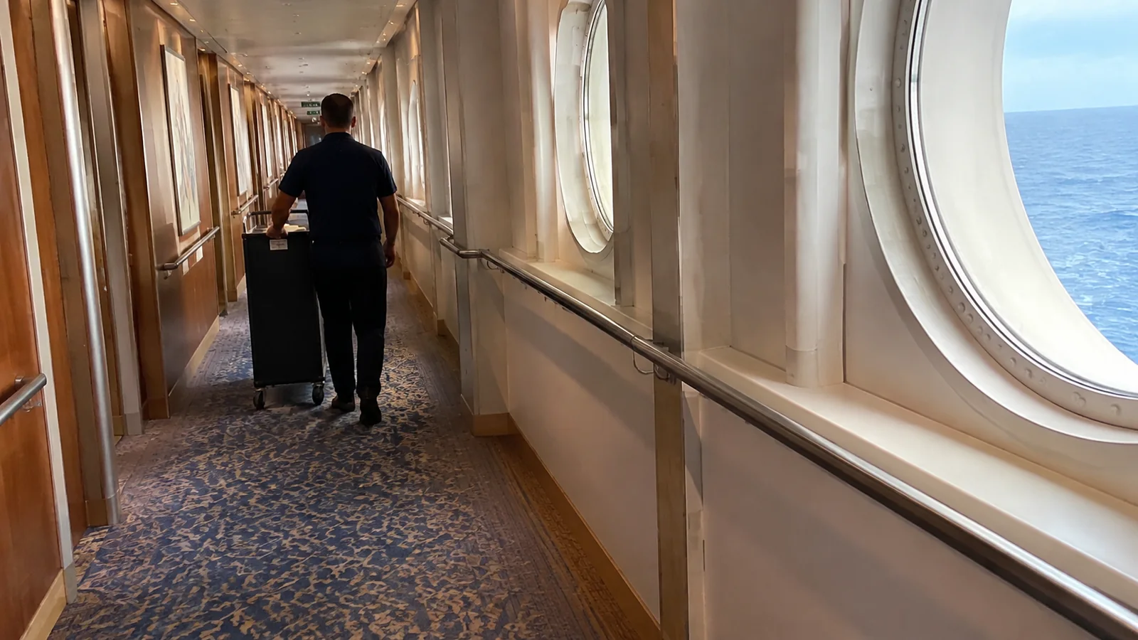 Crew member pushes a cart along a corridor with round windows.