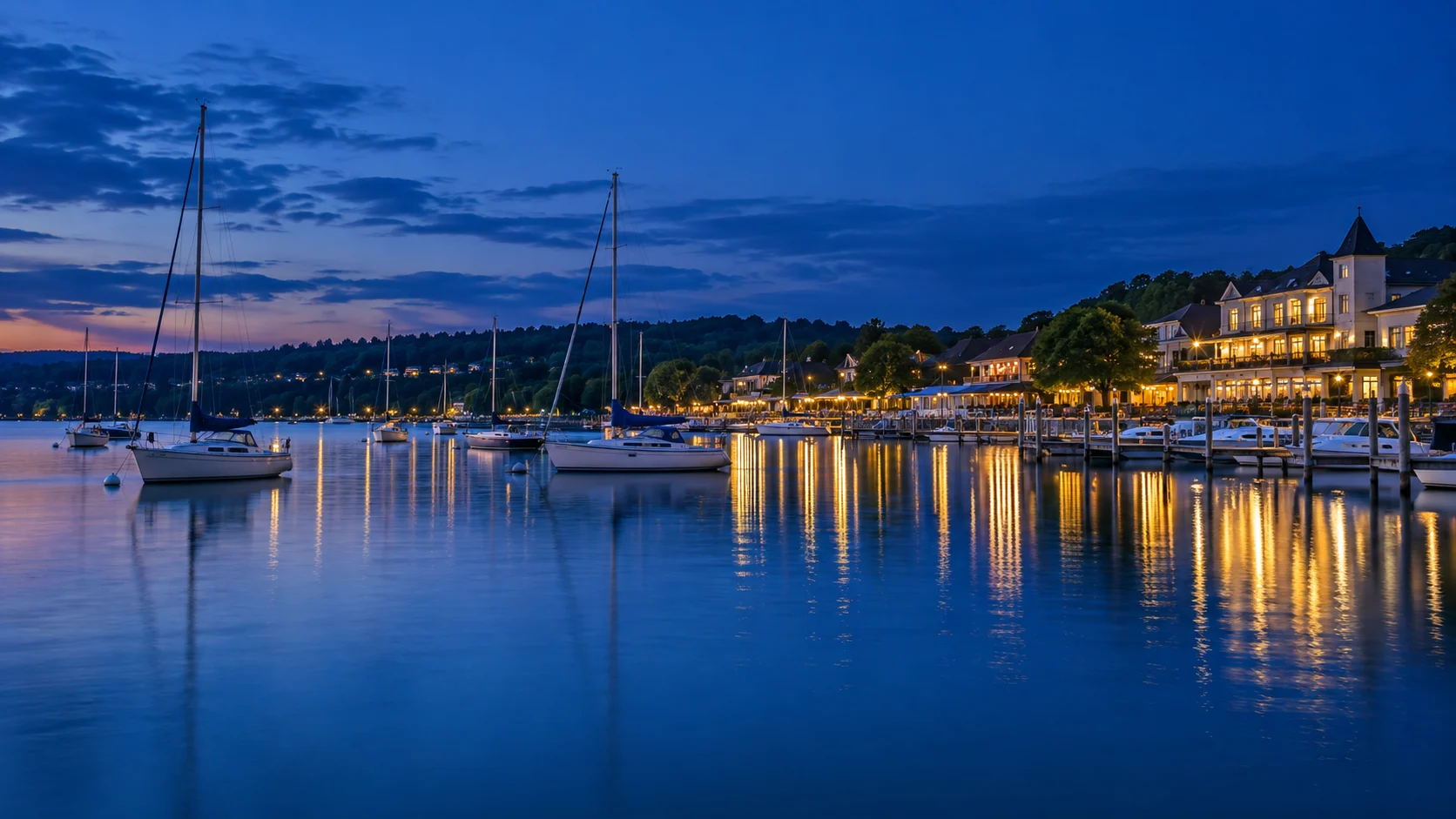 Sailboats rest in Lake Geneva's blue-hour harbor as warm waterfront lights reflect on the water.