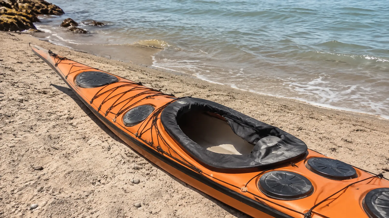 Orange sea kayak rests on sand beside gentle waves.