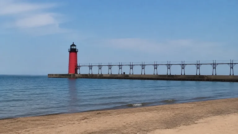 Red lighthouse stands at the end of a long pier over blue water.