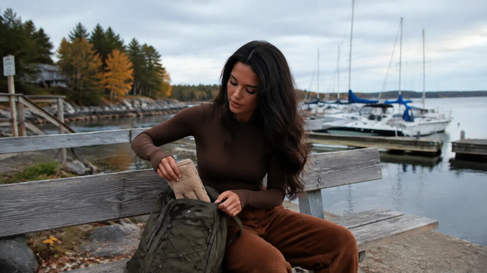 Woman packing a backpack on a marina bench beside sailboats.