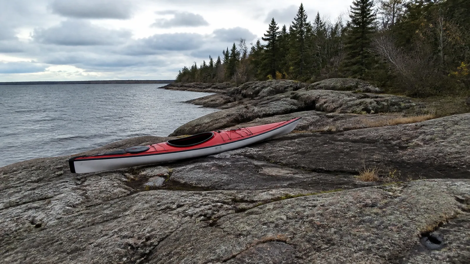 Red kayak rests on flat rocks beside a wooded lakeshore.