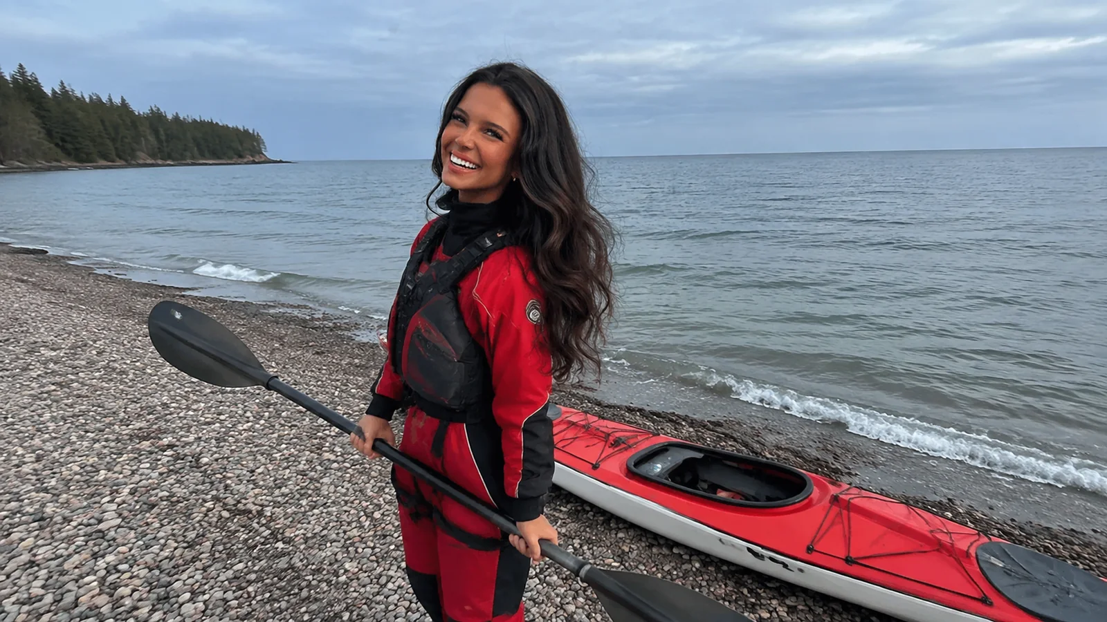 Woman holds a paddle beside a red kayak on a pebble beach.