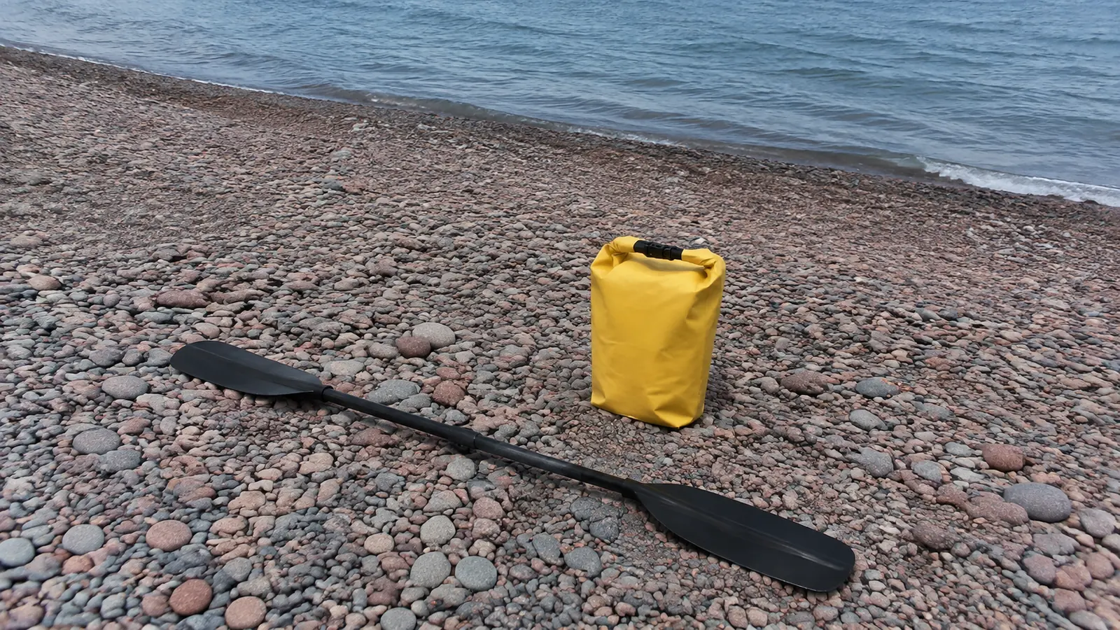 Yellow dry bag and paddle rest on rounded shoreline stones.