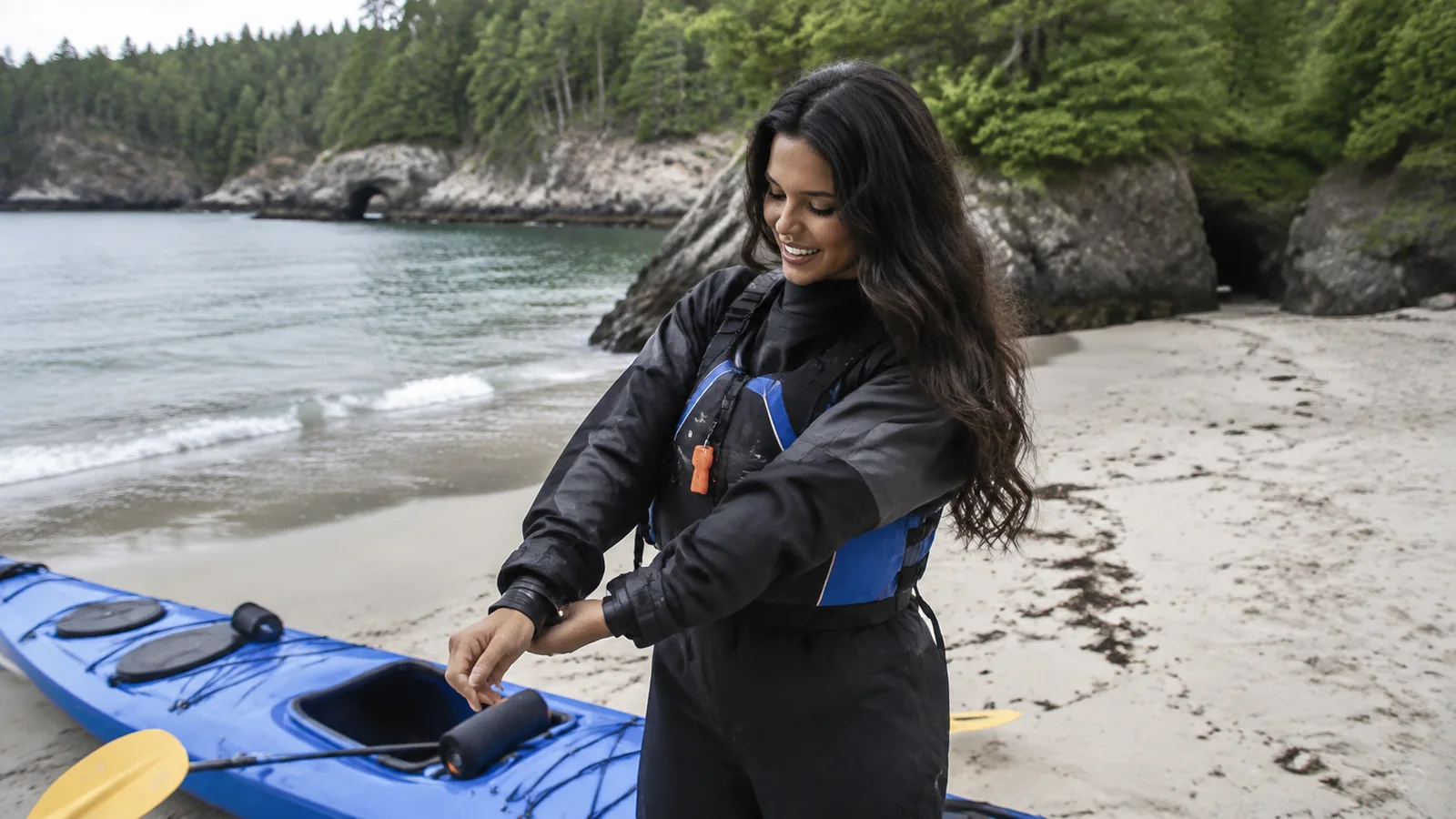 Woman adjusts her sleeve beside a blue kayak on a sandy beach.