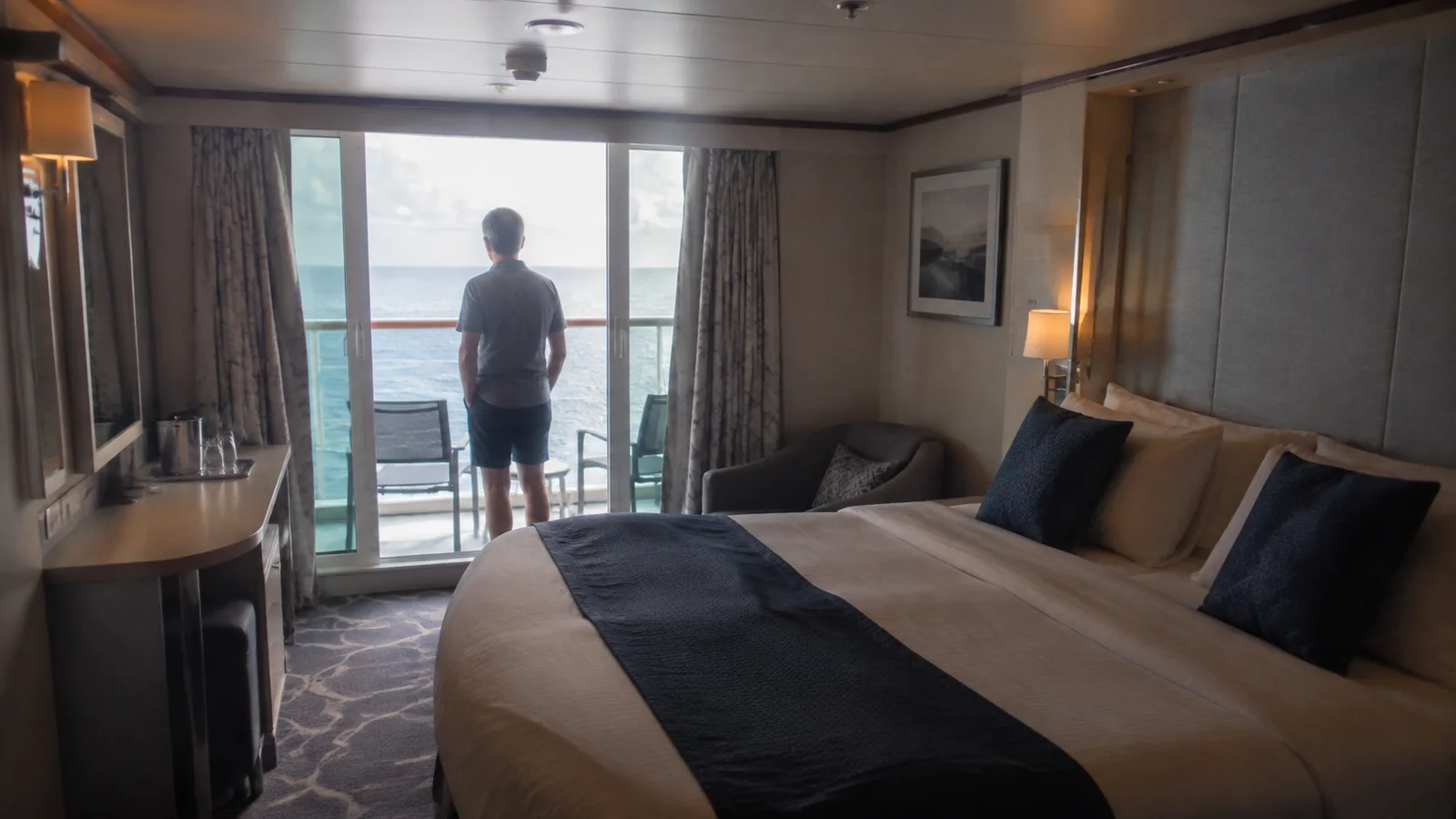 Stateroom bed faces an open balcony doorway where a man looks out to sea.