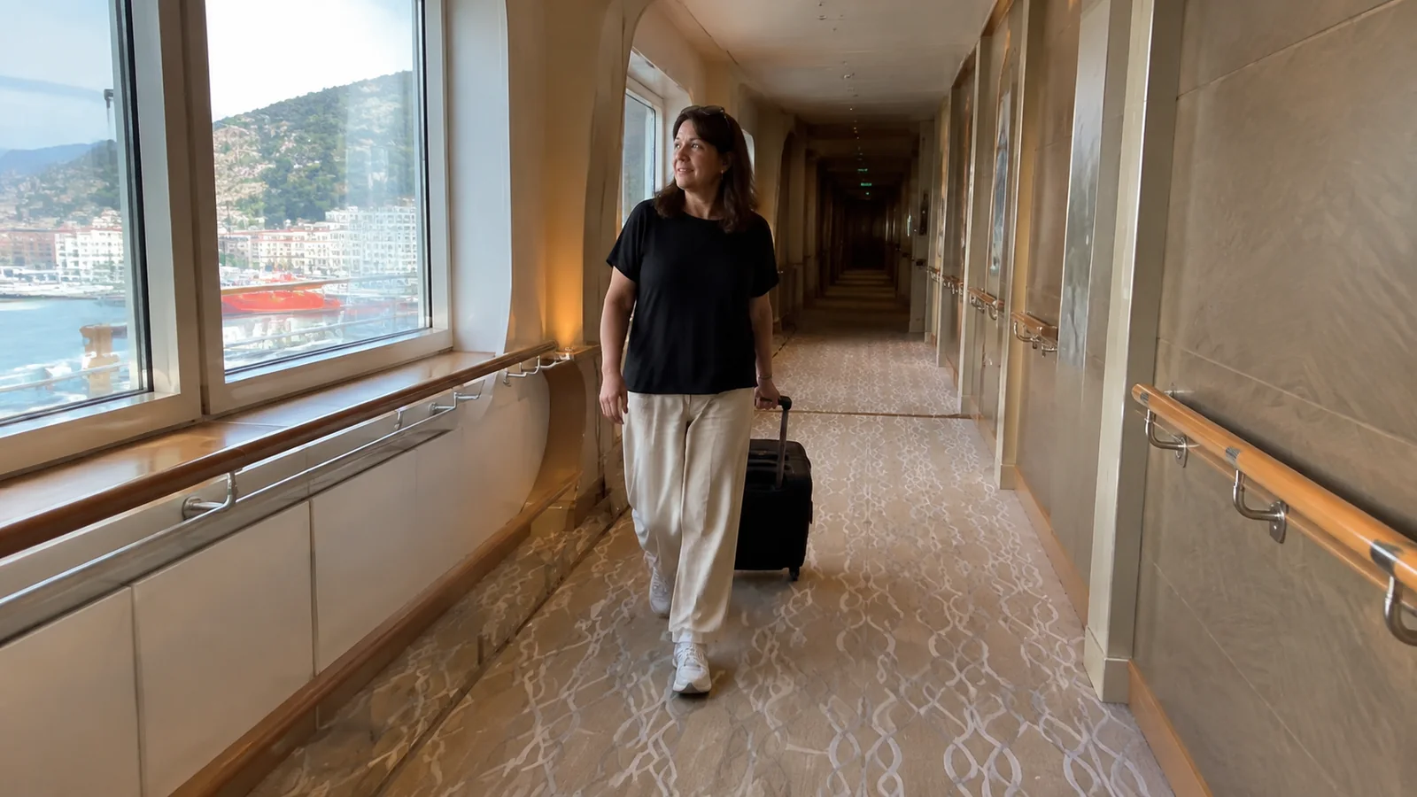 Woman pulls a suitcase through a ship corridor lined with large windows.