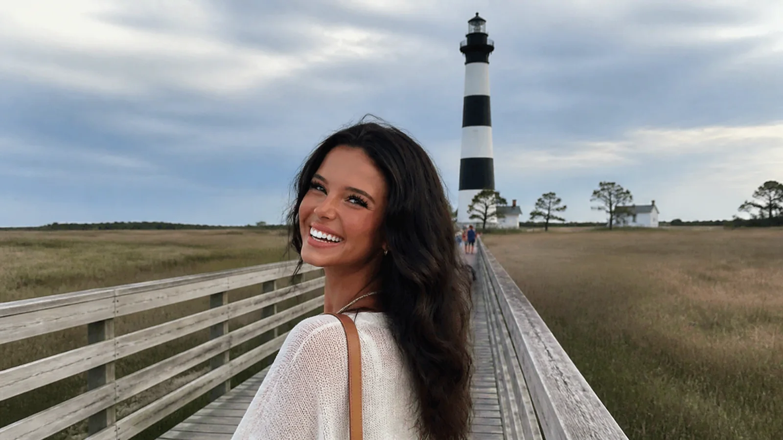 Smiling woman on a boardwalk leading toward a black-and-white striped lighthouse