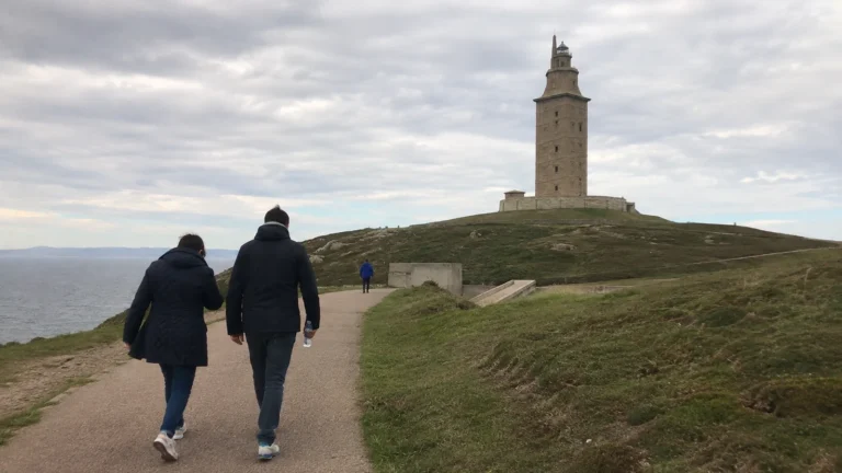 Visitors walk toward a stone lighthouse on a grassy coastal headland.