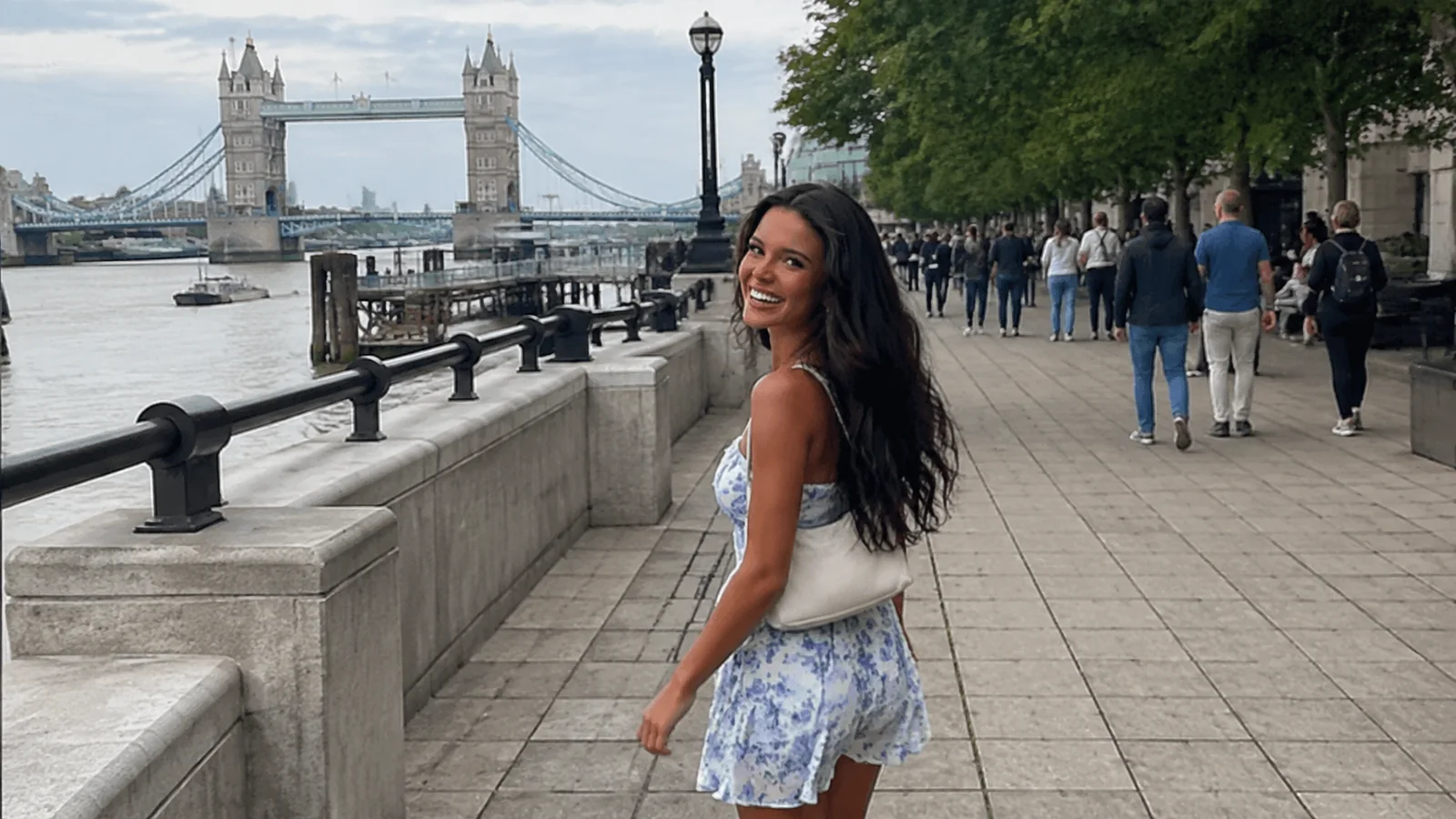 Woman walking a busy river promenade near a twin-towered bridge.