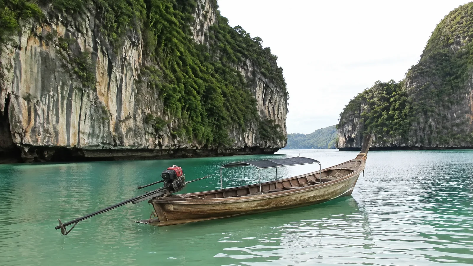 Wooden long-tail boat floats between steep limestone cliffs in green water.