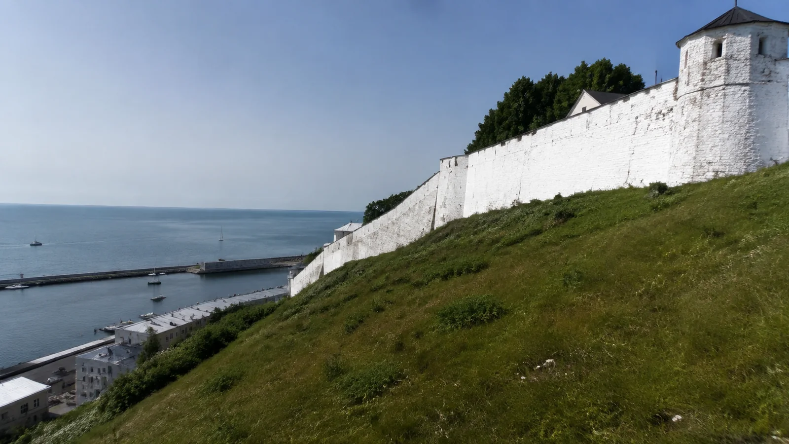 White fort walls rise above a grassy slope overlooking blue water.