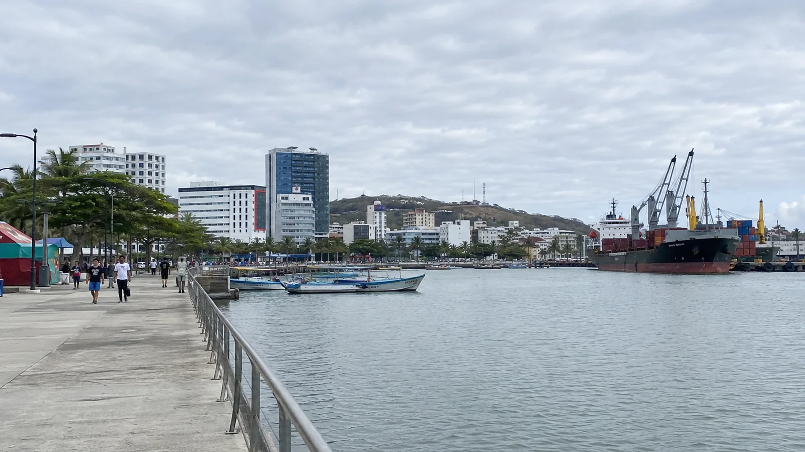 Waterfront promenade with small boats, buildings and port cranes.