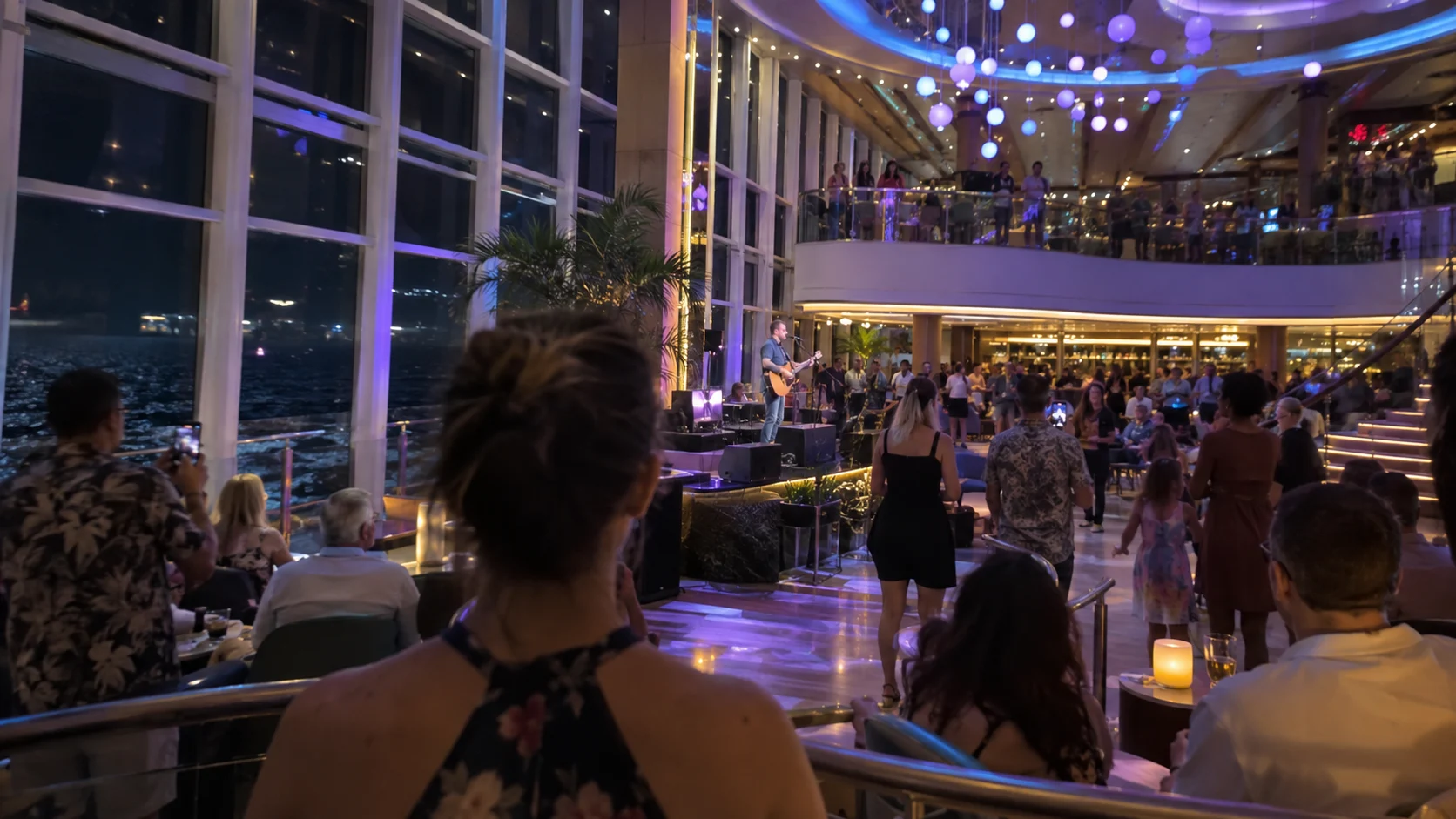 Passengers watching live music in a glass-walled cruise ship atrium at night.