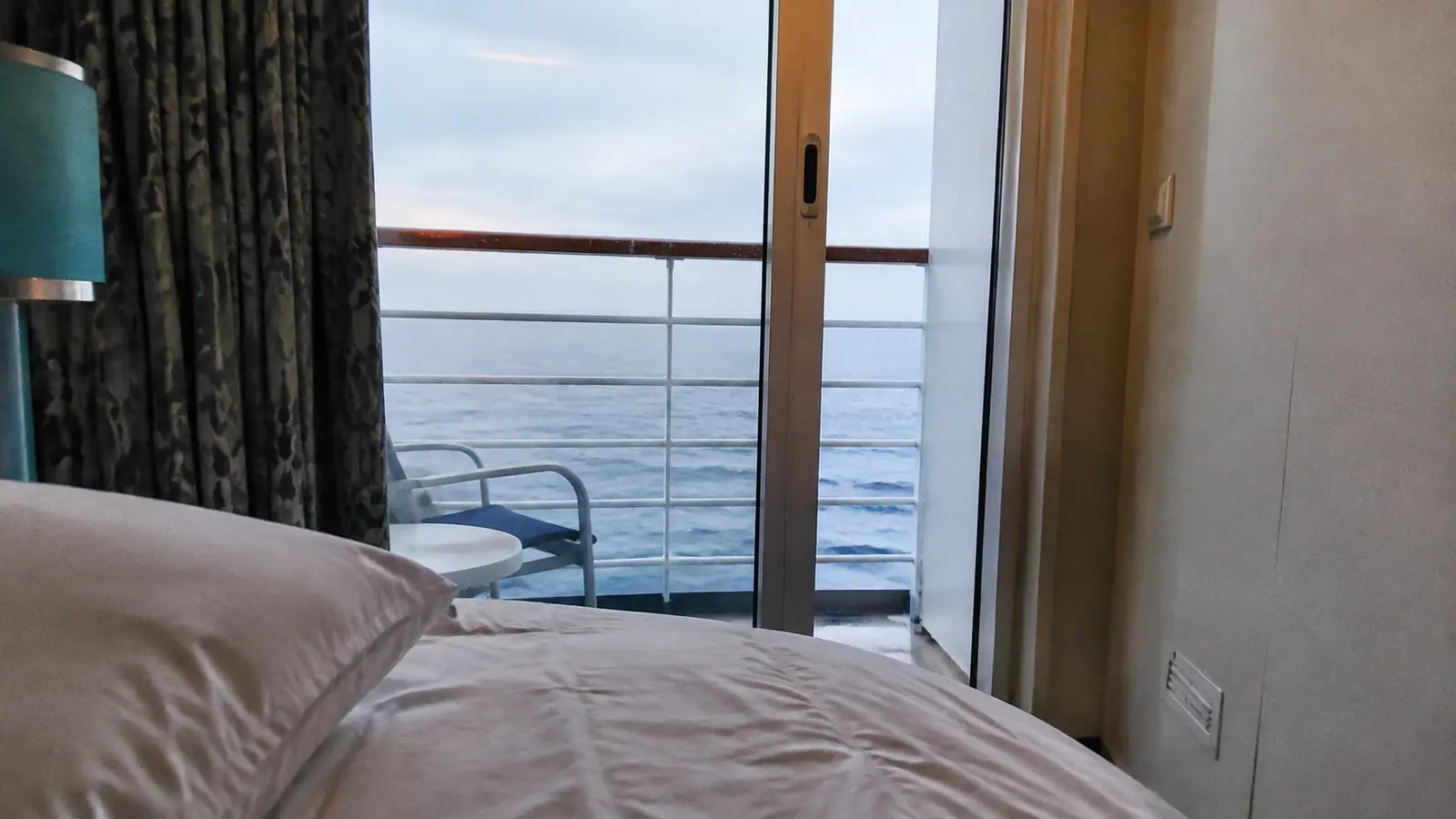 Cruise cabin bed beside sliding balcony doors overlooking the ocean