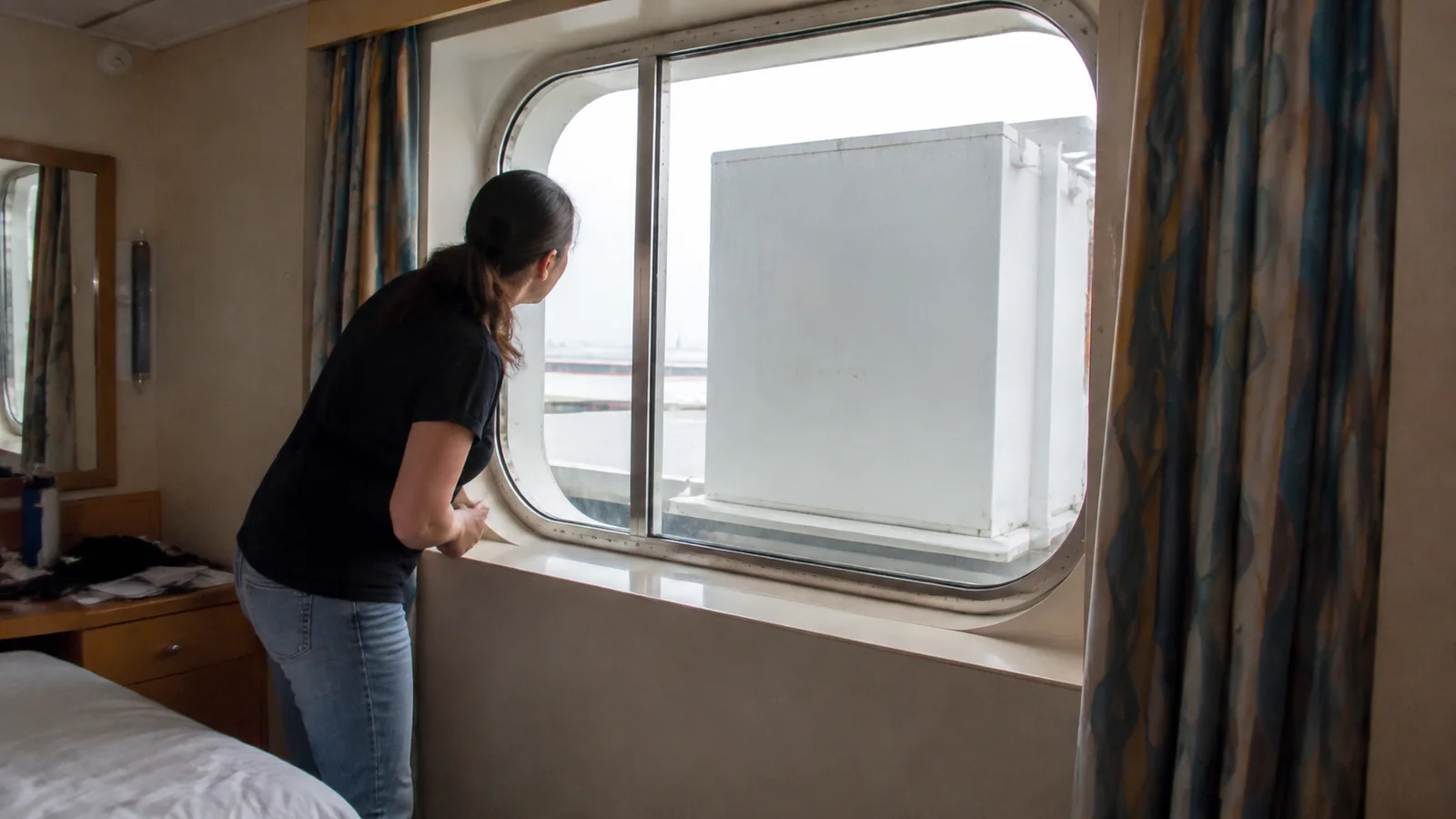 Woman looking through a cruise cabin window partly blocked by an exterior panel