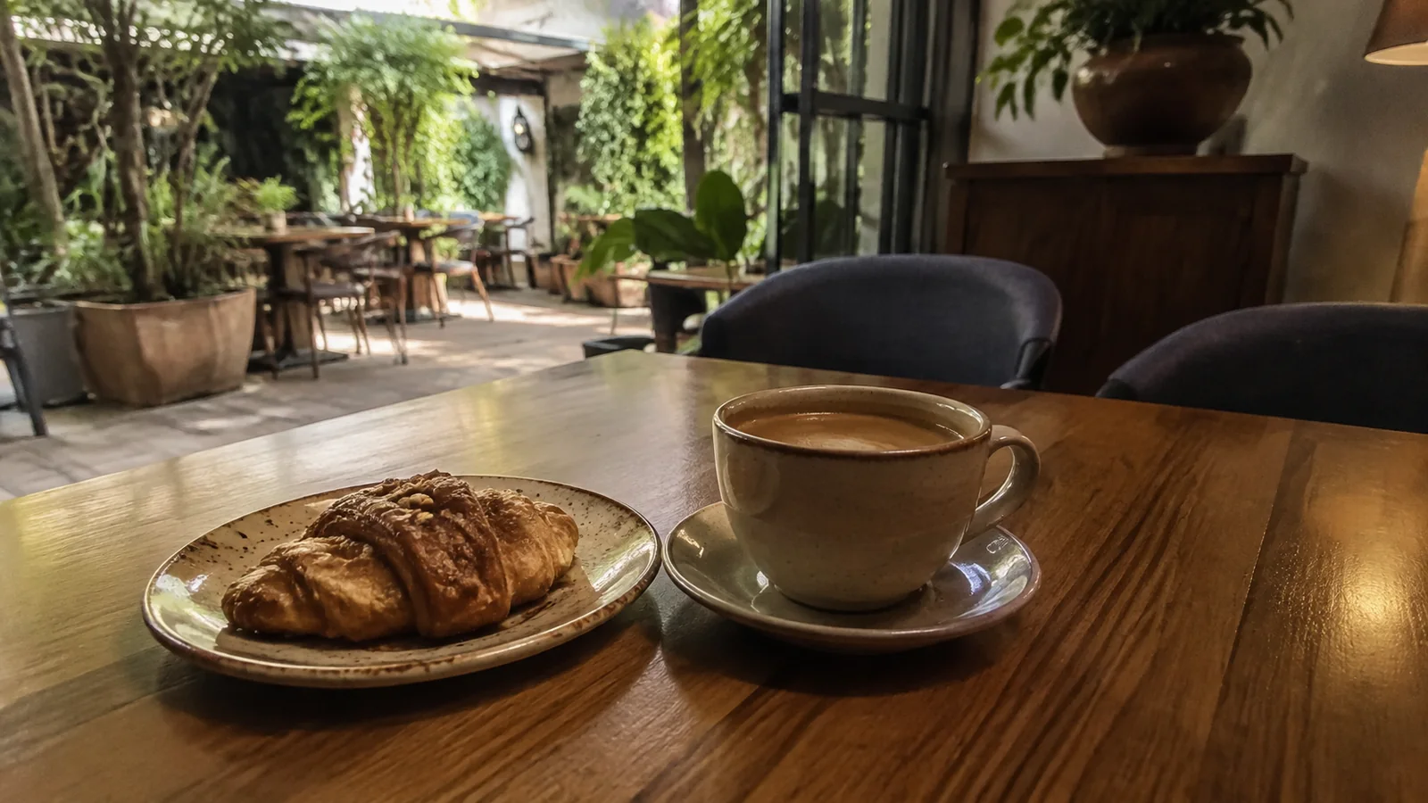 Coffee and a croissant on a wooden table in a leafy cafe.