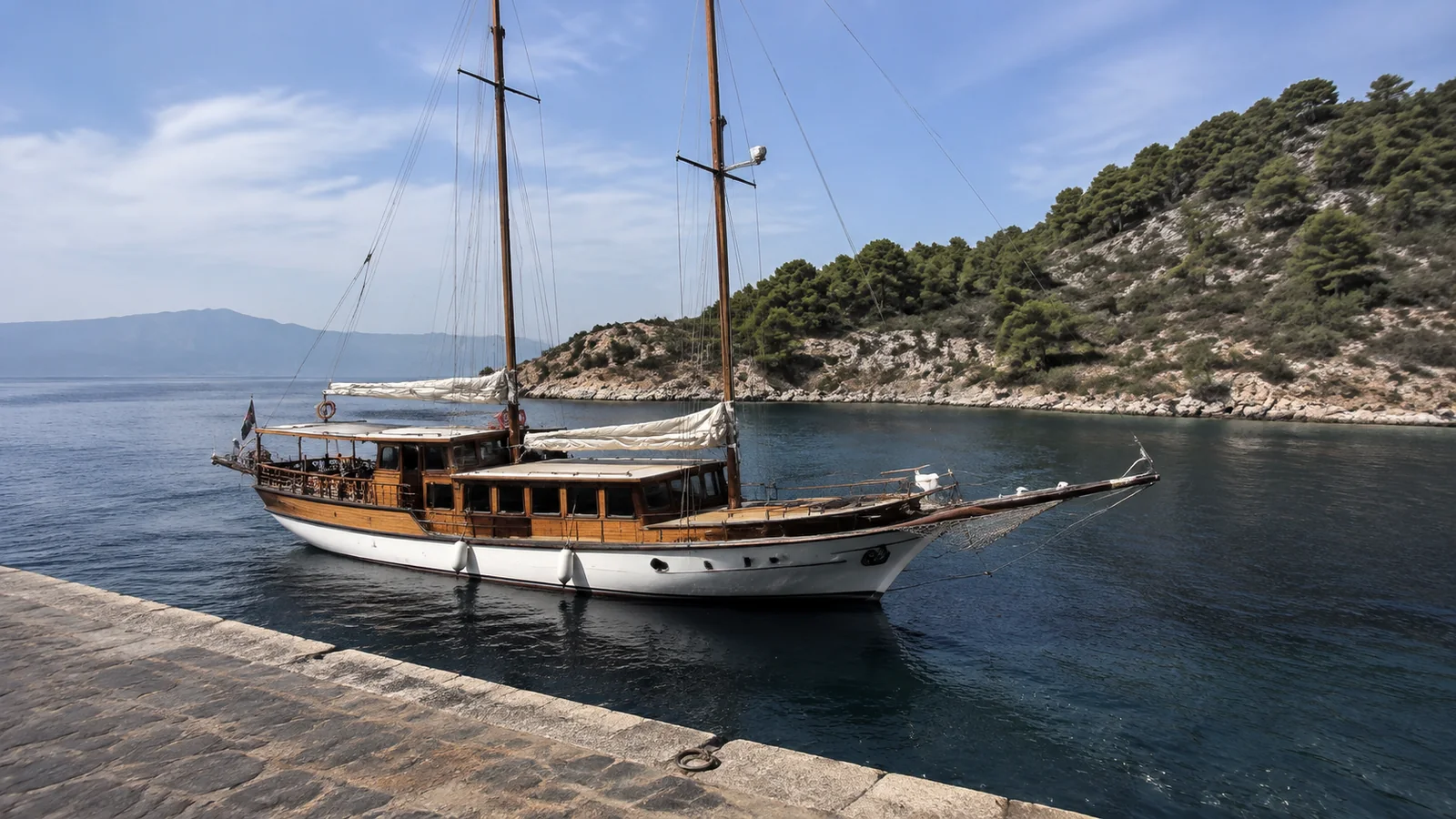 Wooden sailing vessel rests beside a quay in a wooded bay.