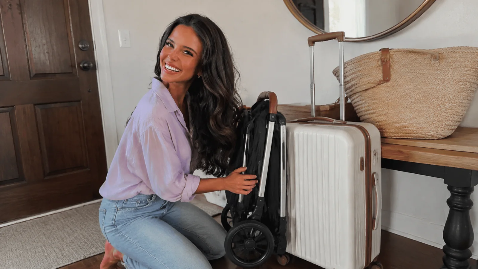 Woman holding a folded stroller beside a suitcase near a doorway.