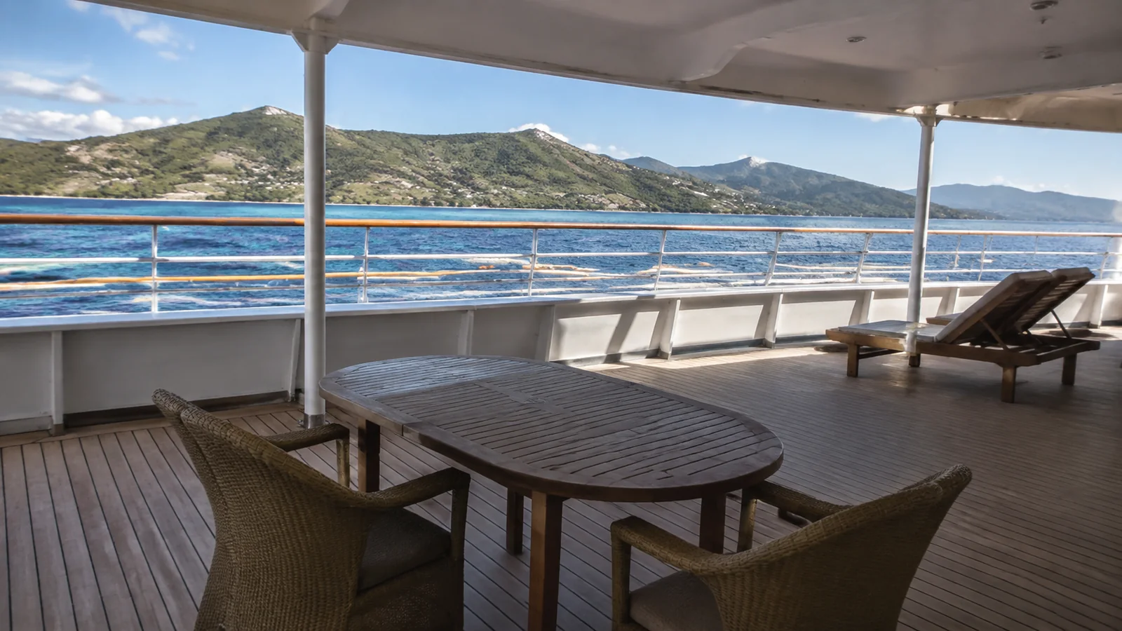 Empty ship deck with a table and chairs facing a mountainous coastline.