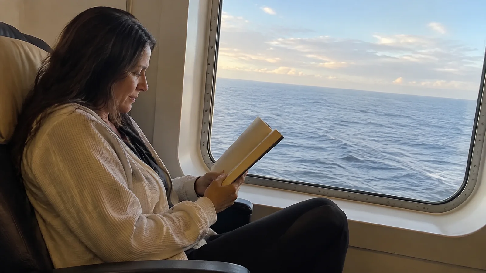 Woman reads a book beside a large window overlooking the sea.
