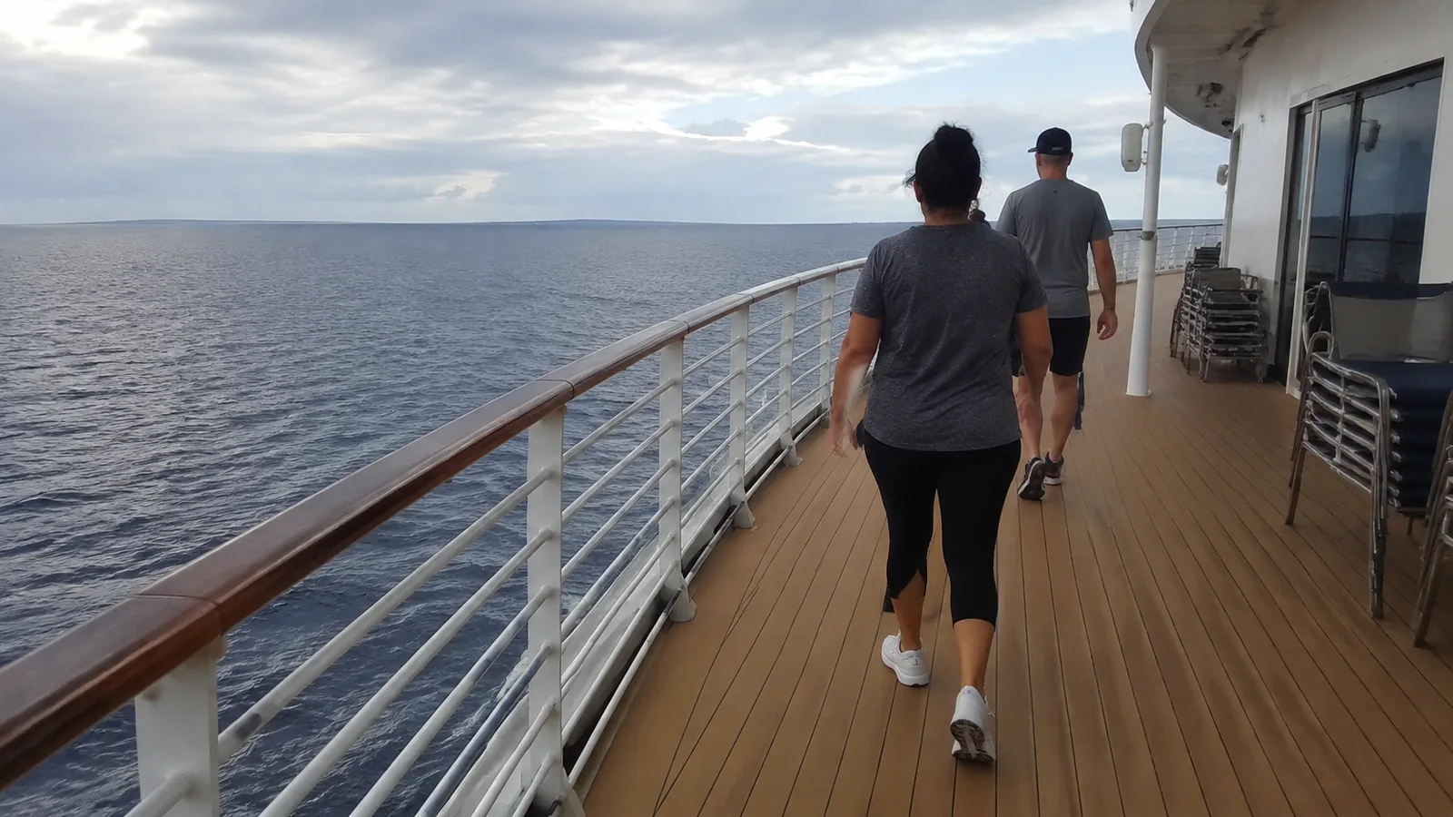 Passengers walk along an outdoor ship deck beside the ocean.
