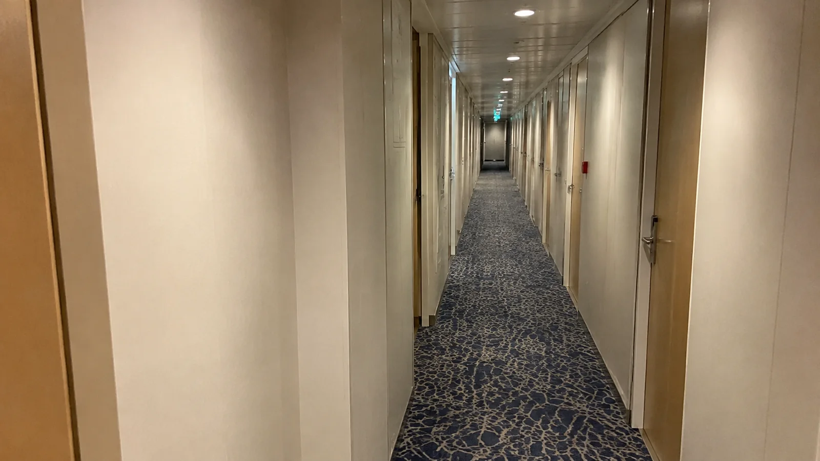Narrow carpeted ship corridor is lined with closed stateroom doors.