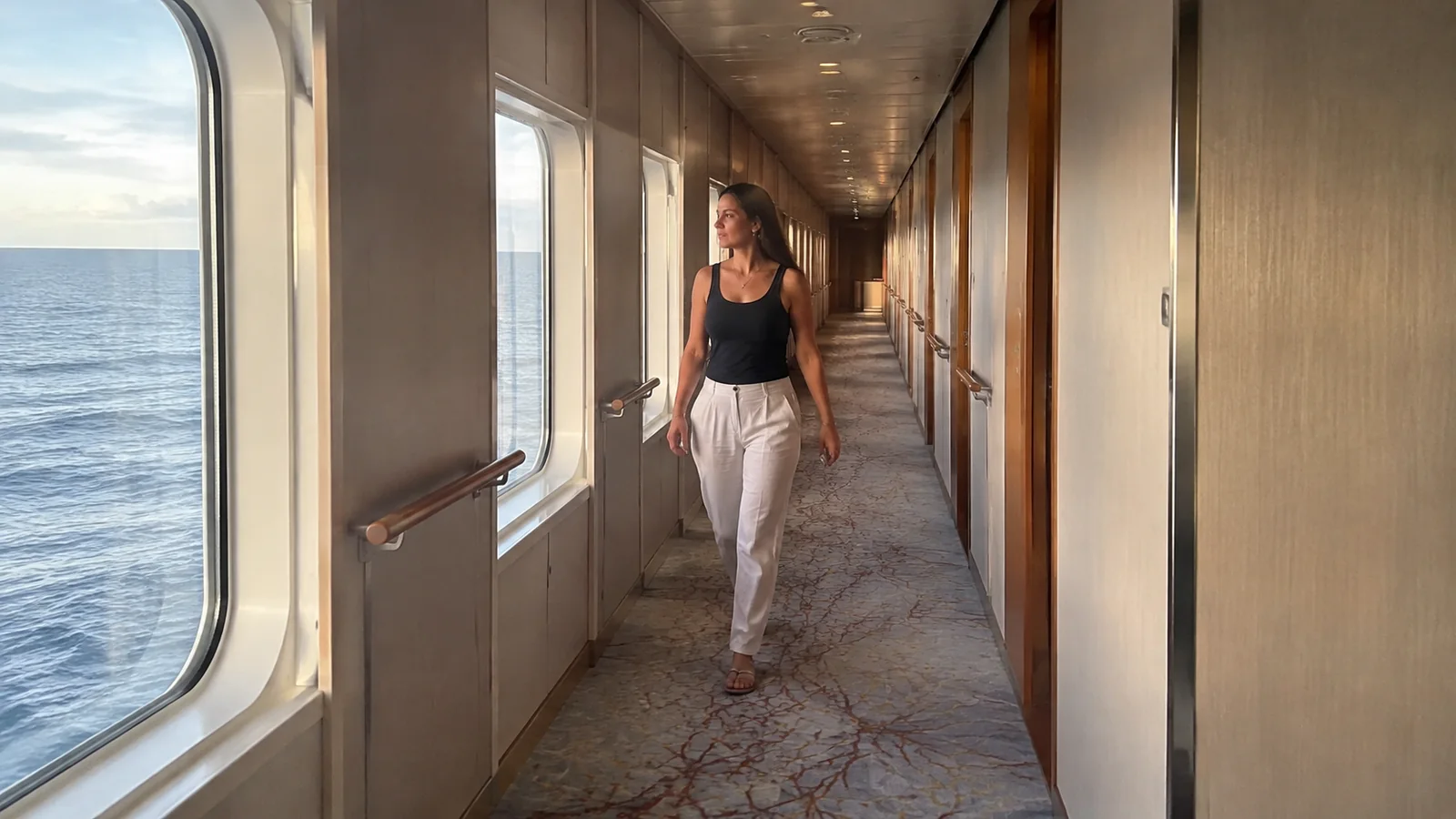 Woman walks along a ship corridor beside ocean windows.