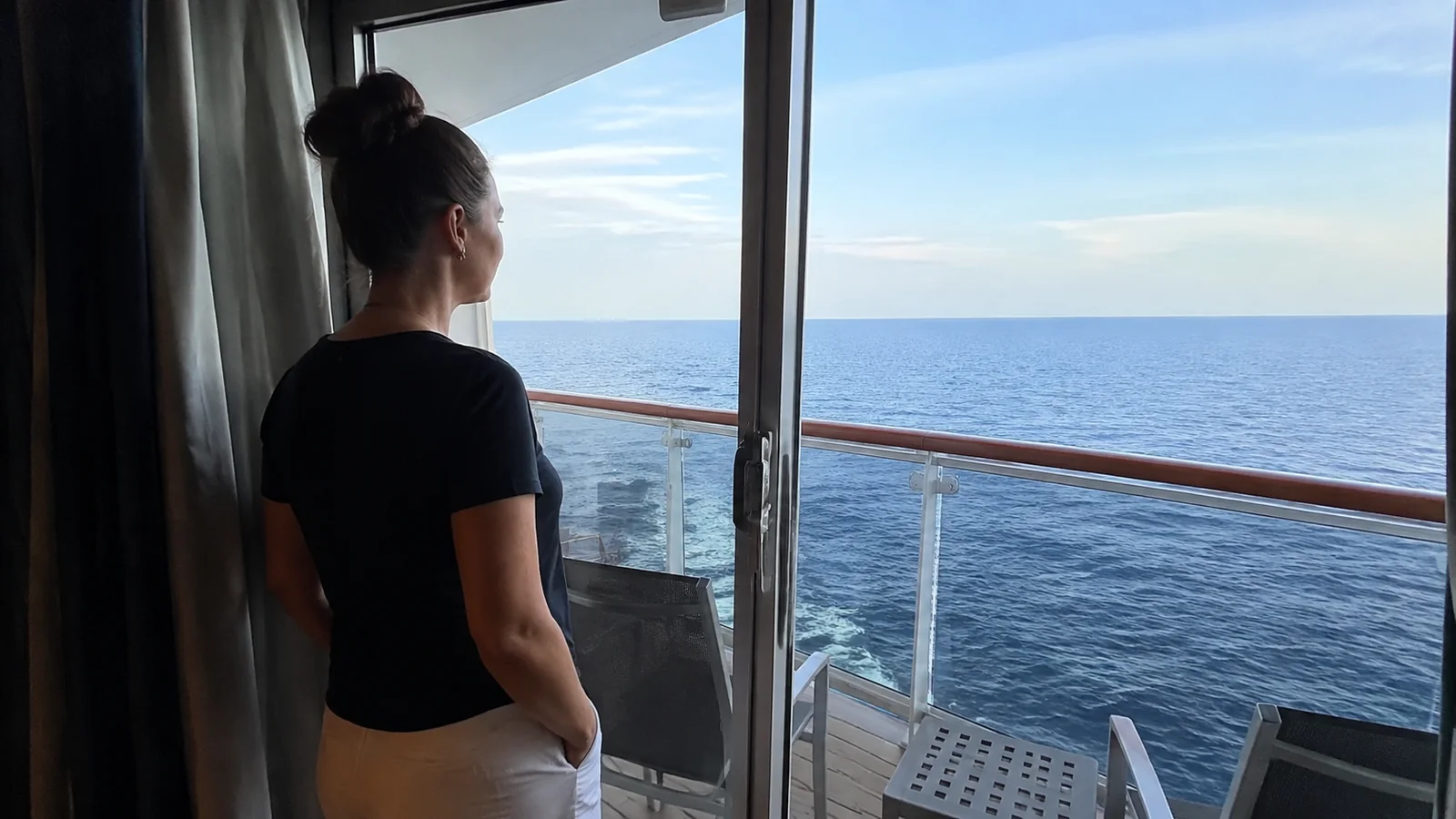 Woman stands beside glass balcony doors looking out to sea.